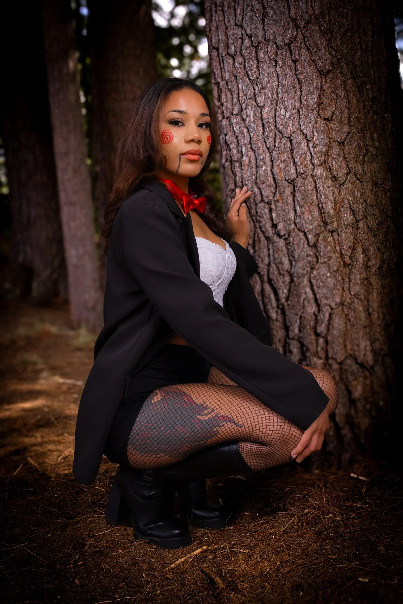 A young woman with makeup and face paint in a forest, wearing a black blazer, white top, fishnet stockings, black boots, and a red bow tie, kneeling next to a tree.