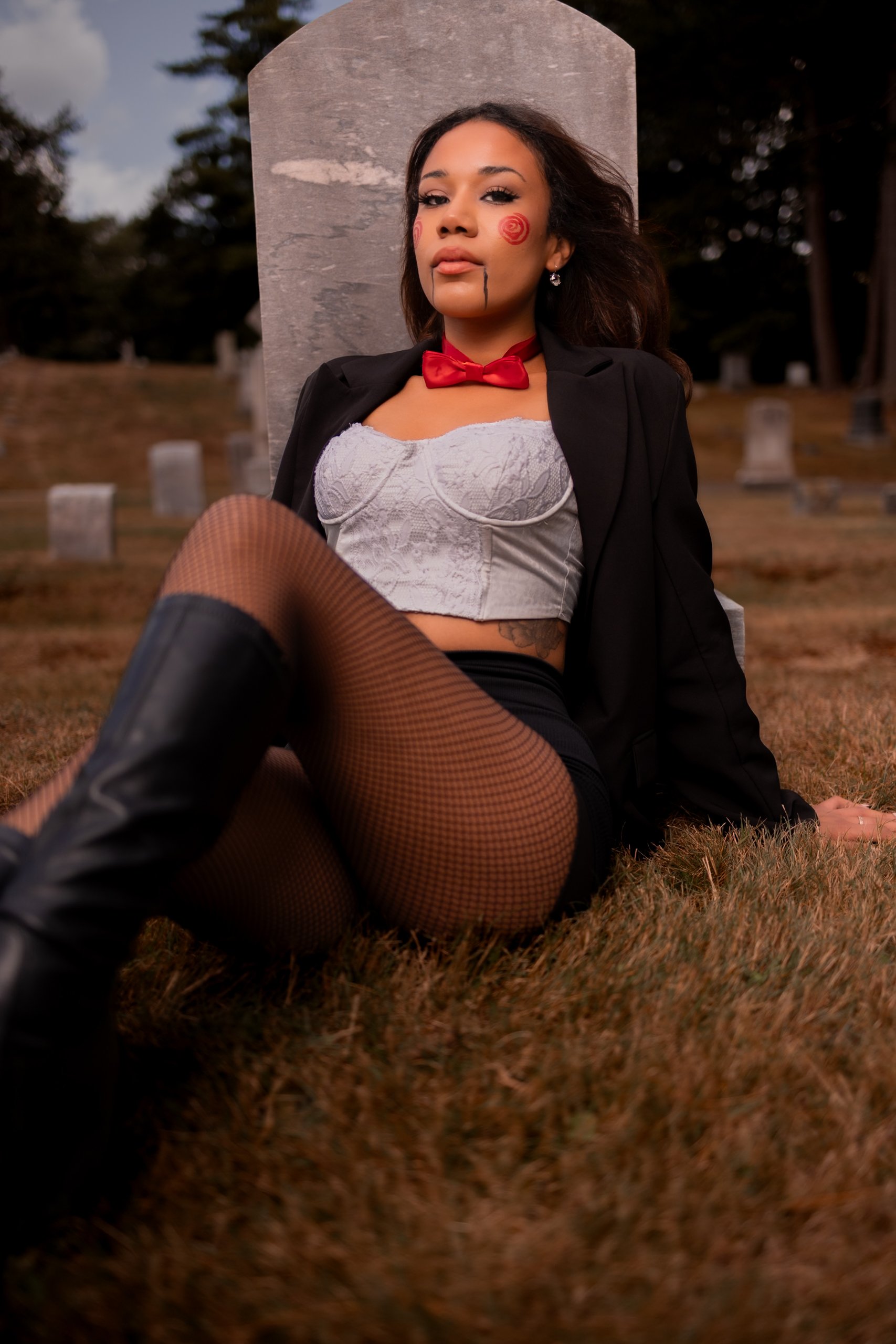 Young woman sitting on grass in costume, with graveyard background, dressed as a clown or mime with face paint, fishnet stockings, boots, and a blazer.