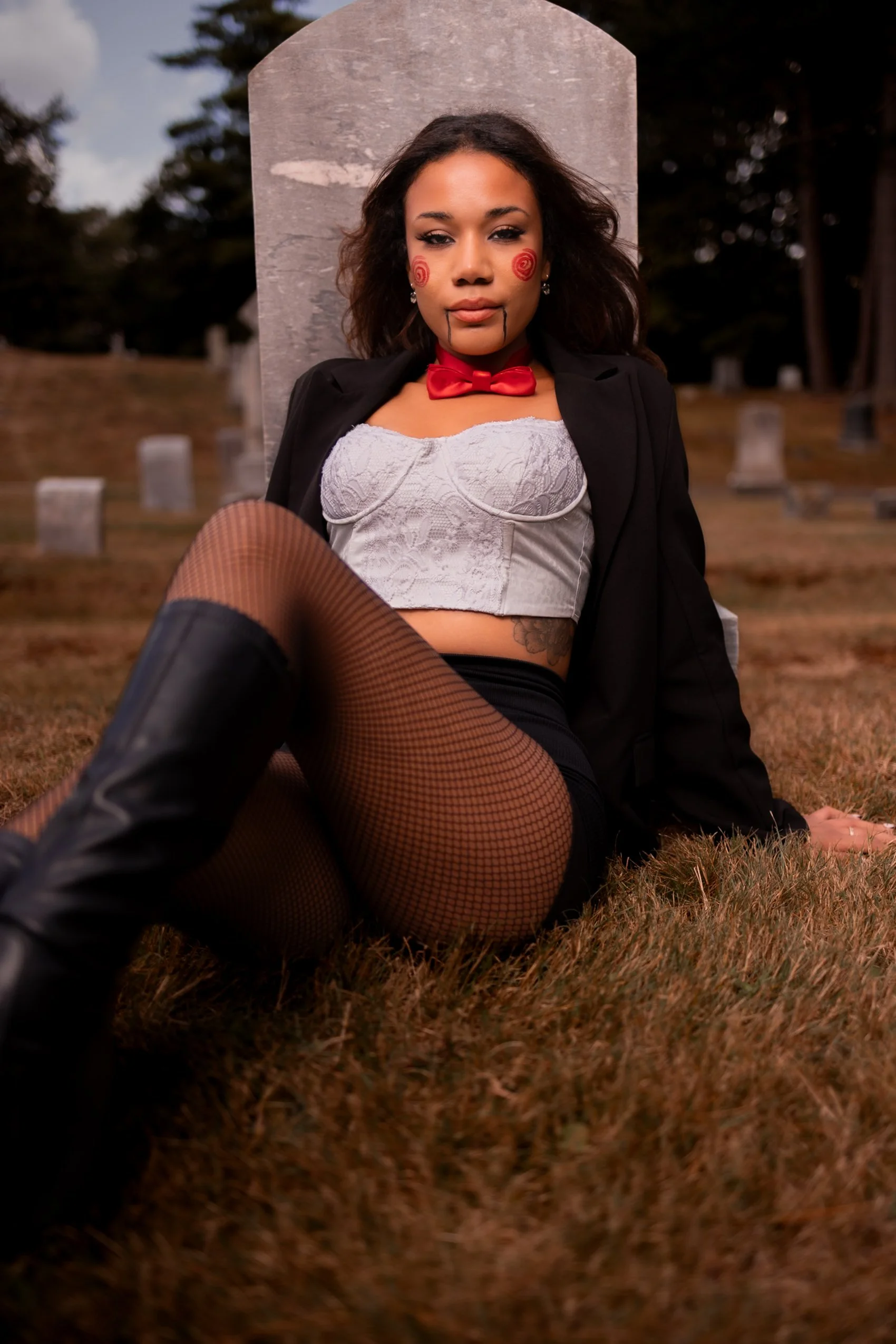 A woman sitting on the grass in a Halloween costume, with a gravestone in the background, wearing makeup that resembles a malevolent doll or clown, with swirl cheeks and black tears, dressed in a lace bustier, black jacket, fishnet stockings, and kne
