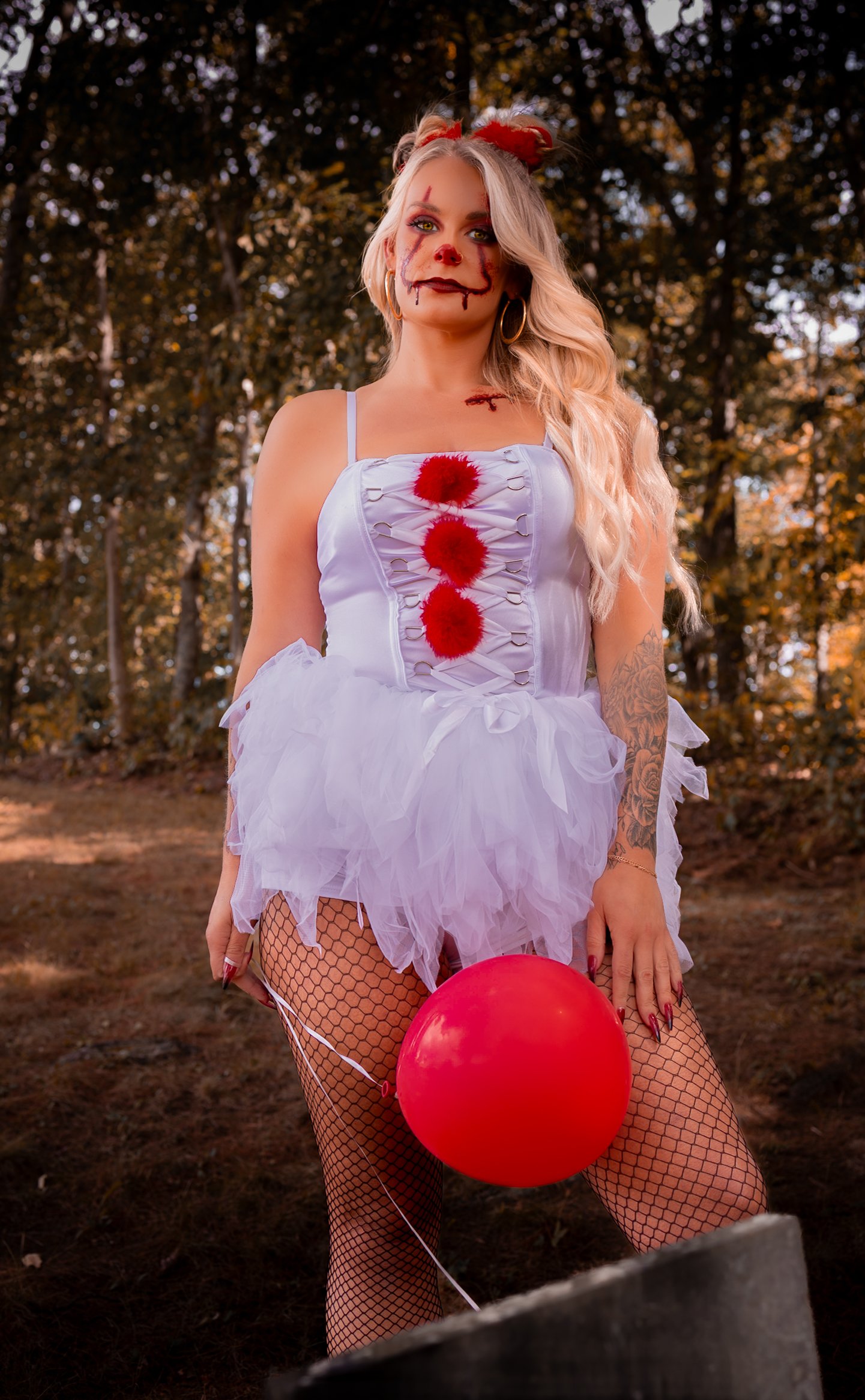Woman dressed as a sinister clown in a white tutu, holding a red balloon, with face paint and theatrical makeup, standing outdoors with trees in the background.