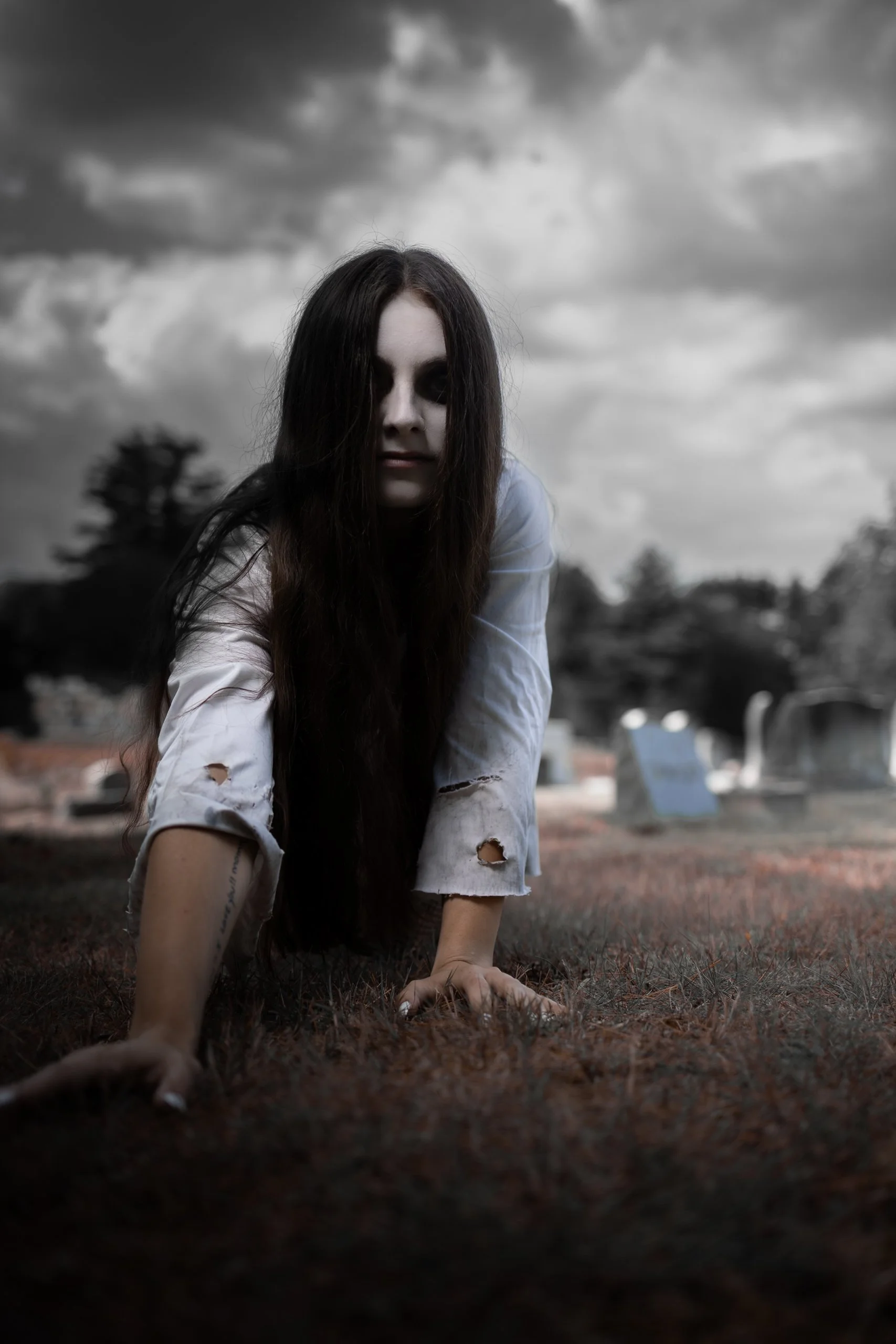 A woman with long dark hair and dark makeup, wearing a torn white shirt, crawling on the ground in a graveyard under a cloudy sky.