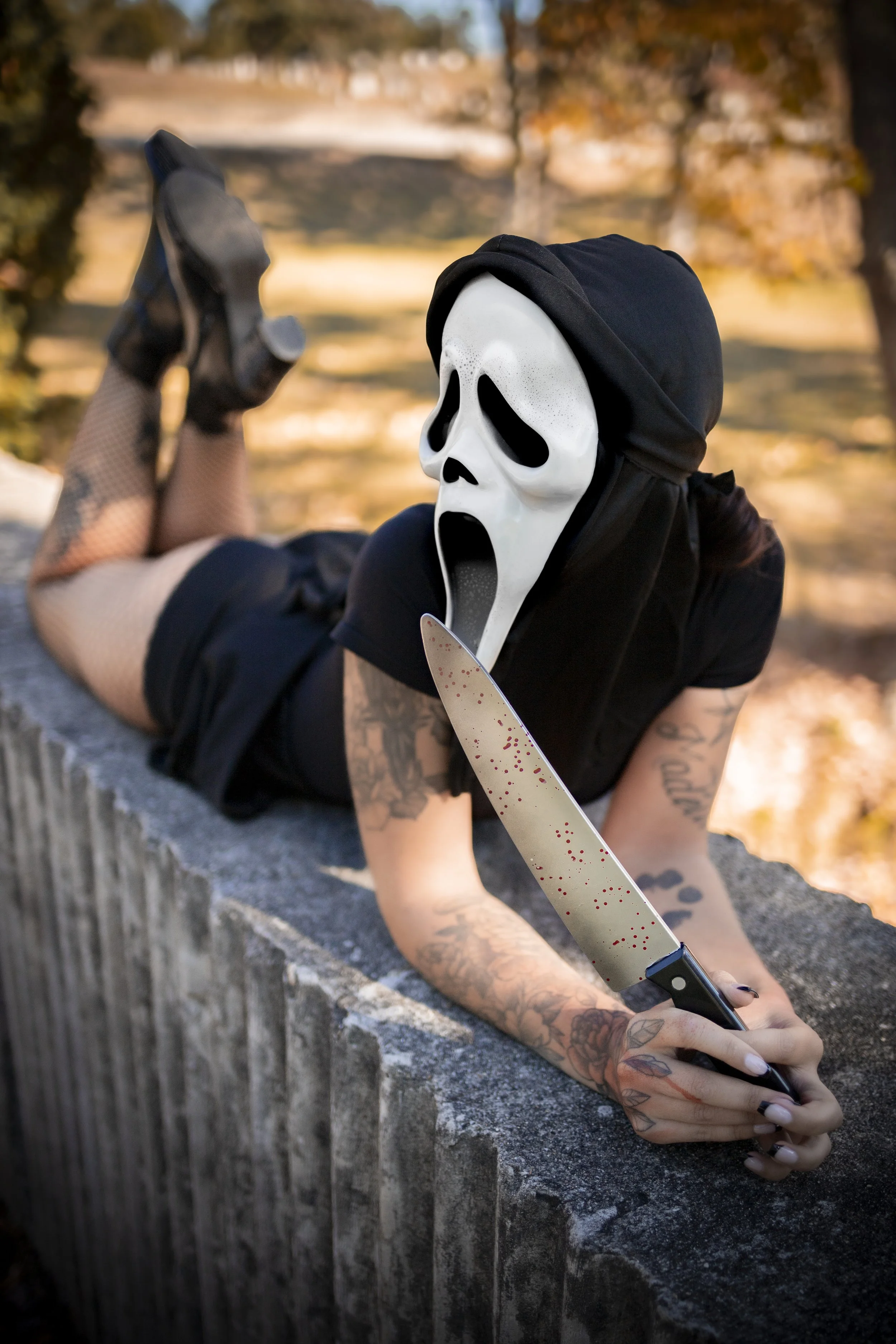 Person wearing a Ghostface mask, black hood, and black outfit lying on a stone ledge outdoors, holding a blood-splattered kitchen knife.