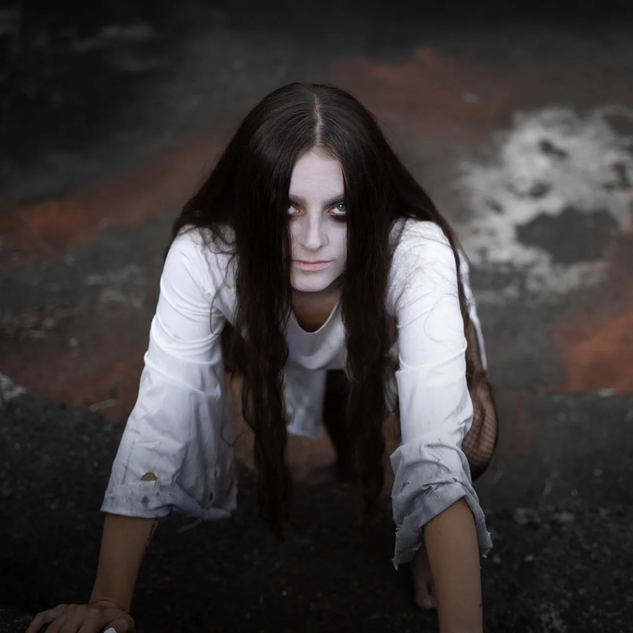 A woman with long dark hair, dressed in white with torn sleeves, crawling on the ground with intense makeup and a sinister expression.