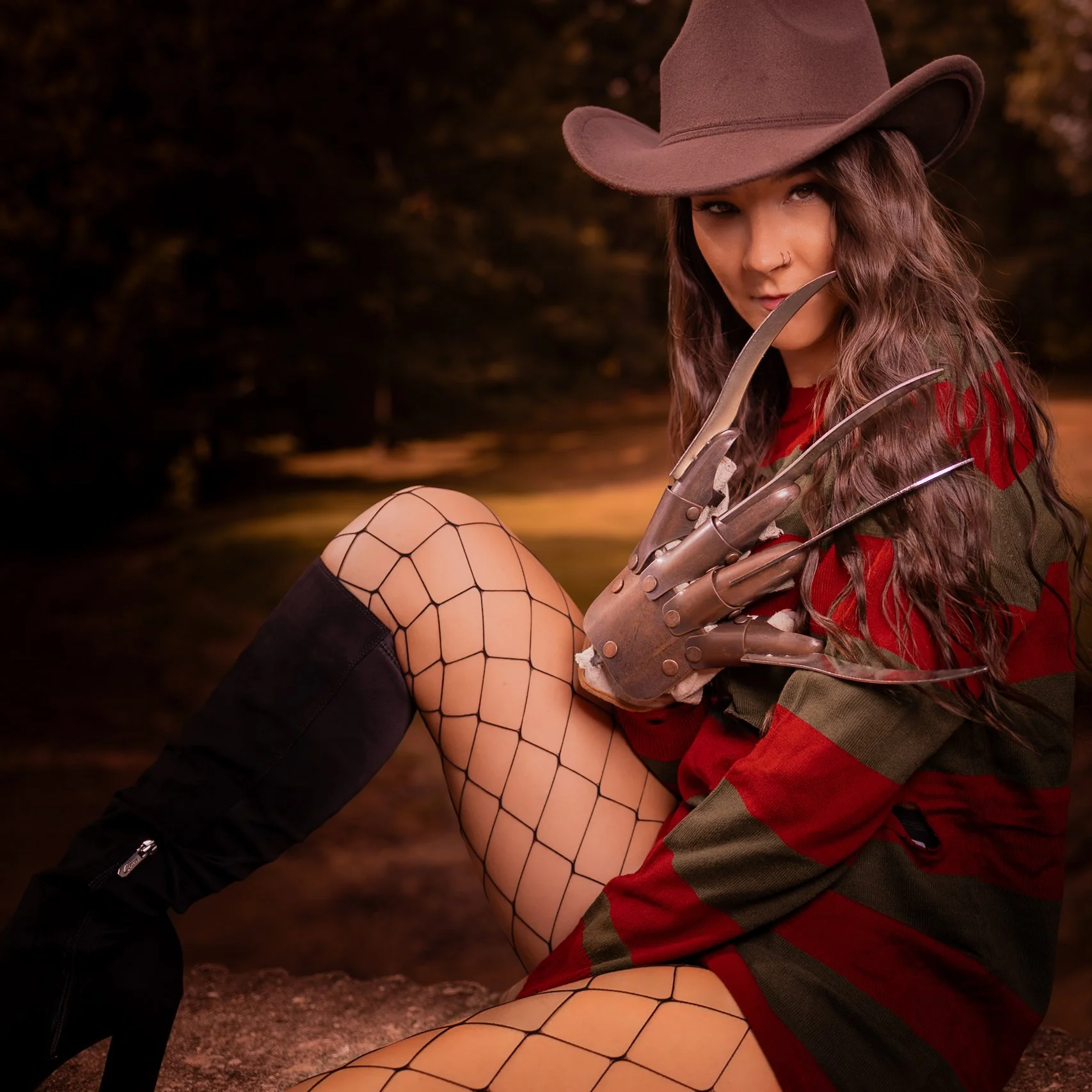 A woman wearing a wide-brimmed hat, fishnet stockings, a military-style jacket, and tall black boots sitting outdoors at sunset with a large metal glove with bladed fingers.