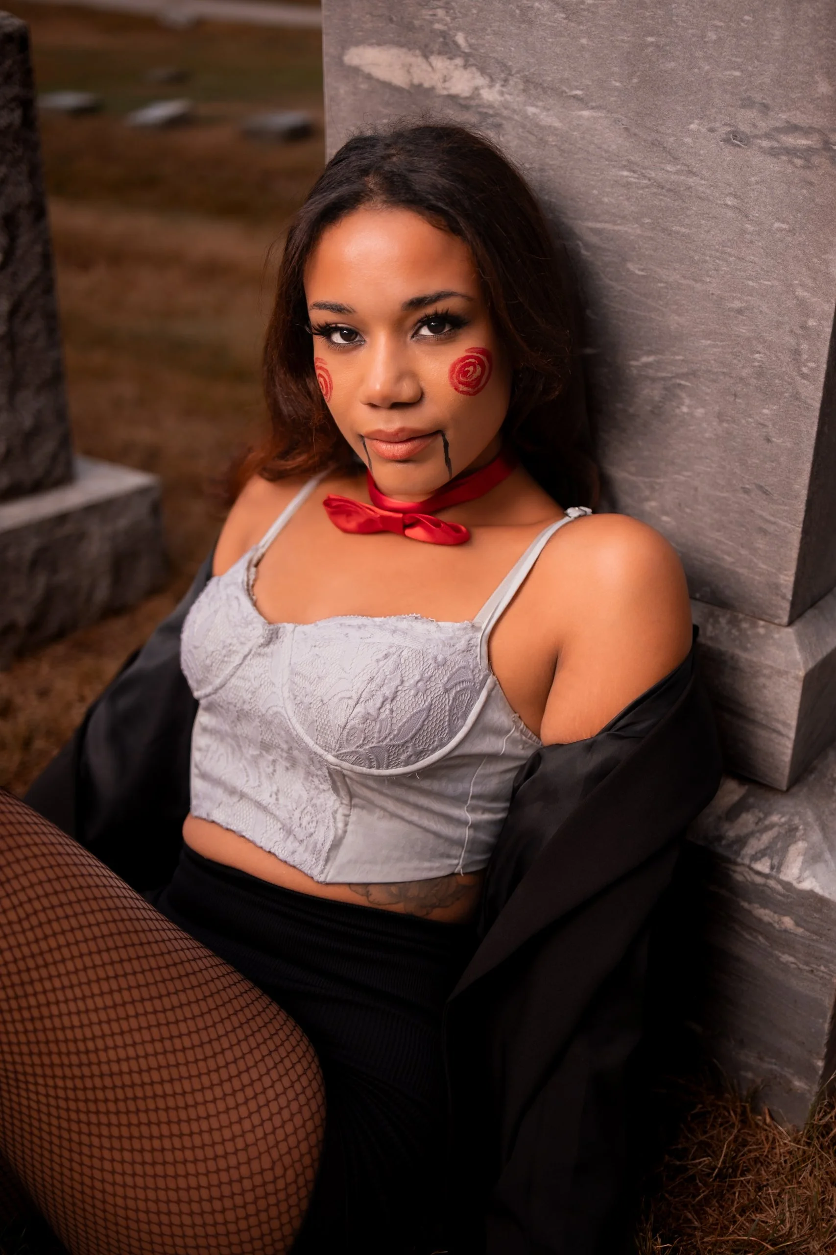 A woman dressed in Halloween costume with face paint resembling a creepy doll, wearing fishnet stockings, a lace crop top, red choker, and black cape, sitting against a stone wall outdoors.