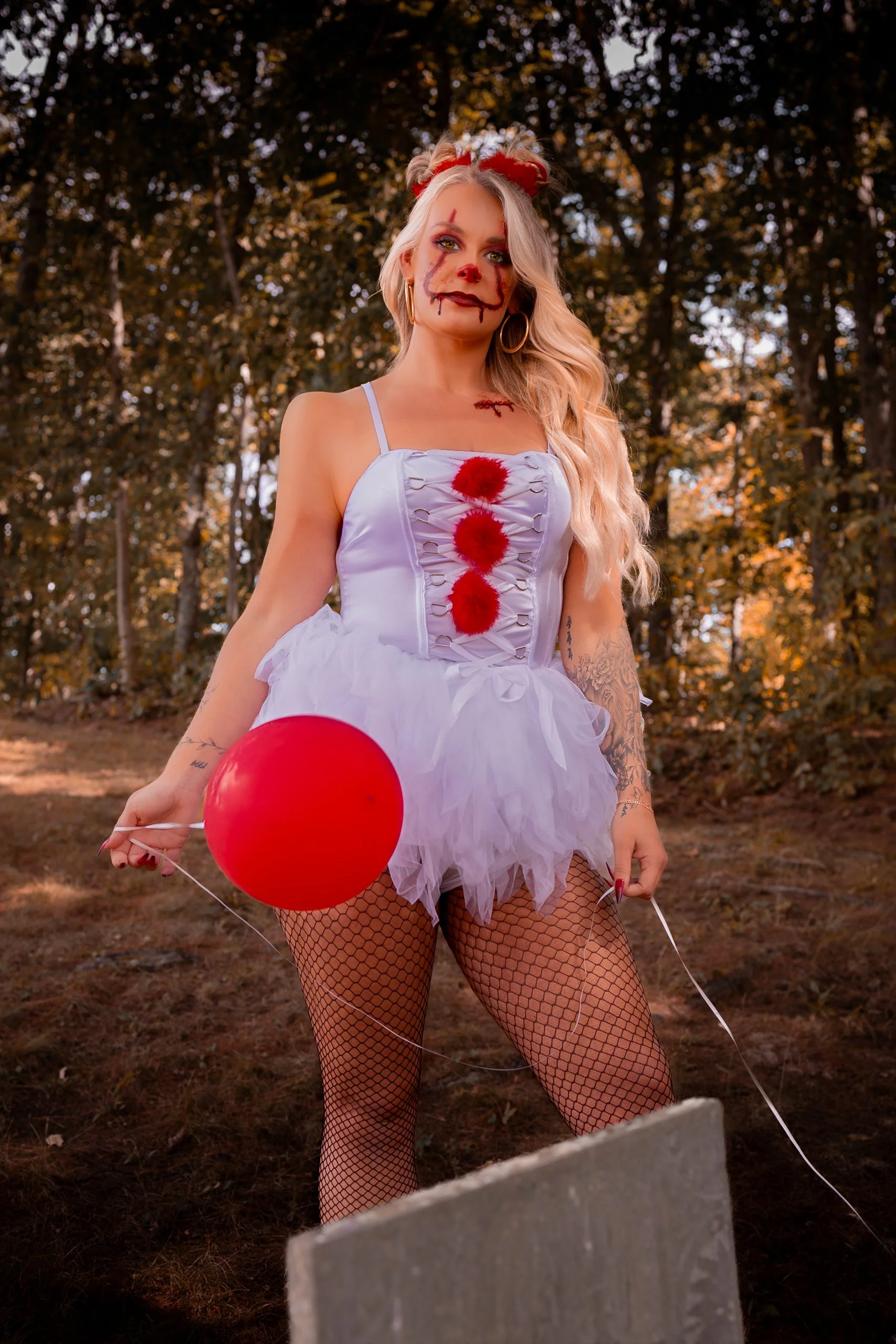 A woman dressed in a clown costume with makeup resembling a creepy clown, holding a red balloon, standing outdoors in a wooded area.
