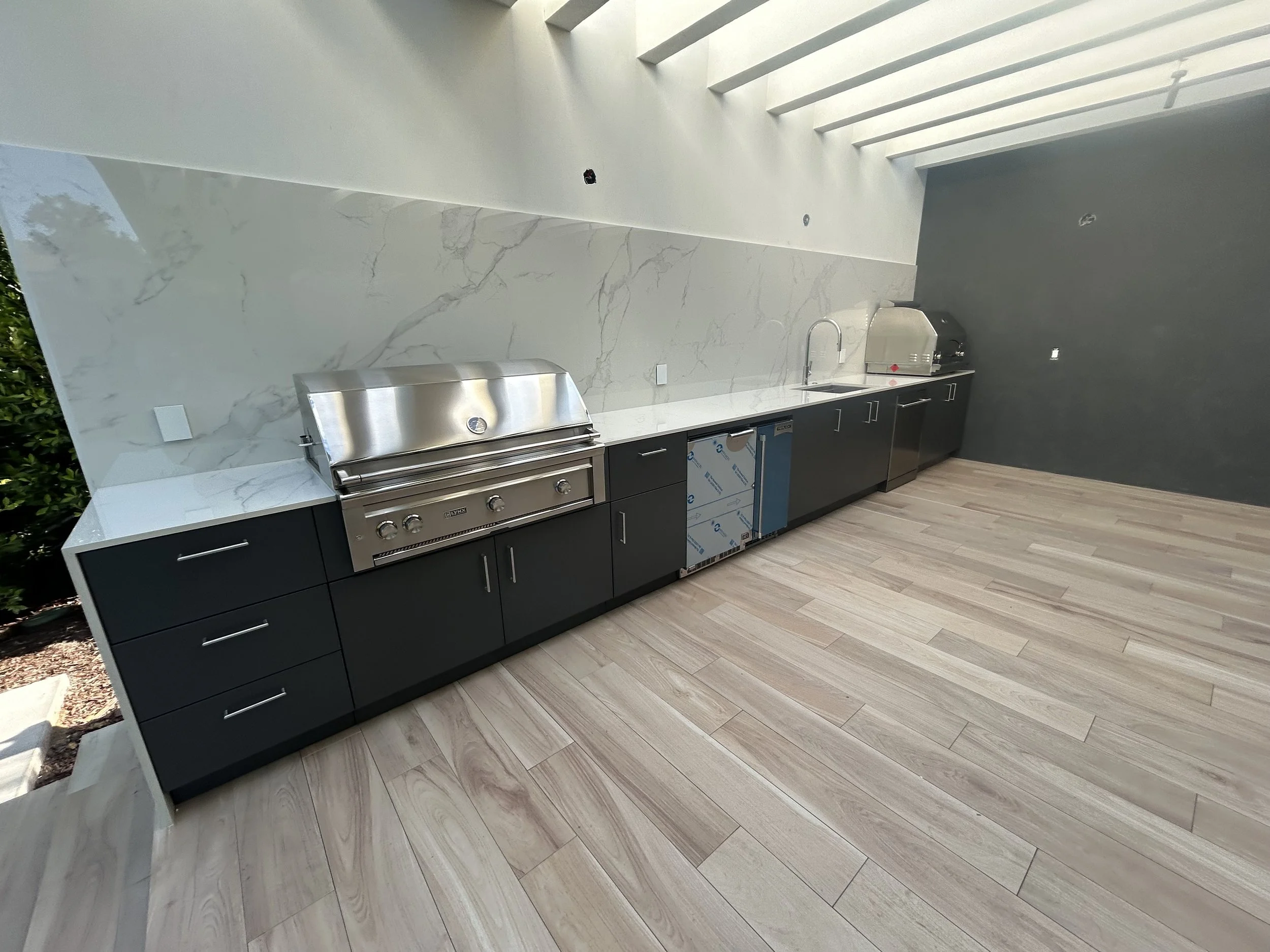 Outdoor kitchen with dark cabinets, a built-in grill, and a stone-look backsplash beneath a covered patio.