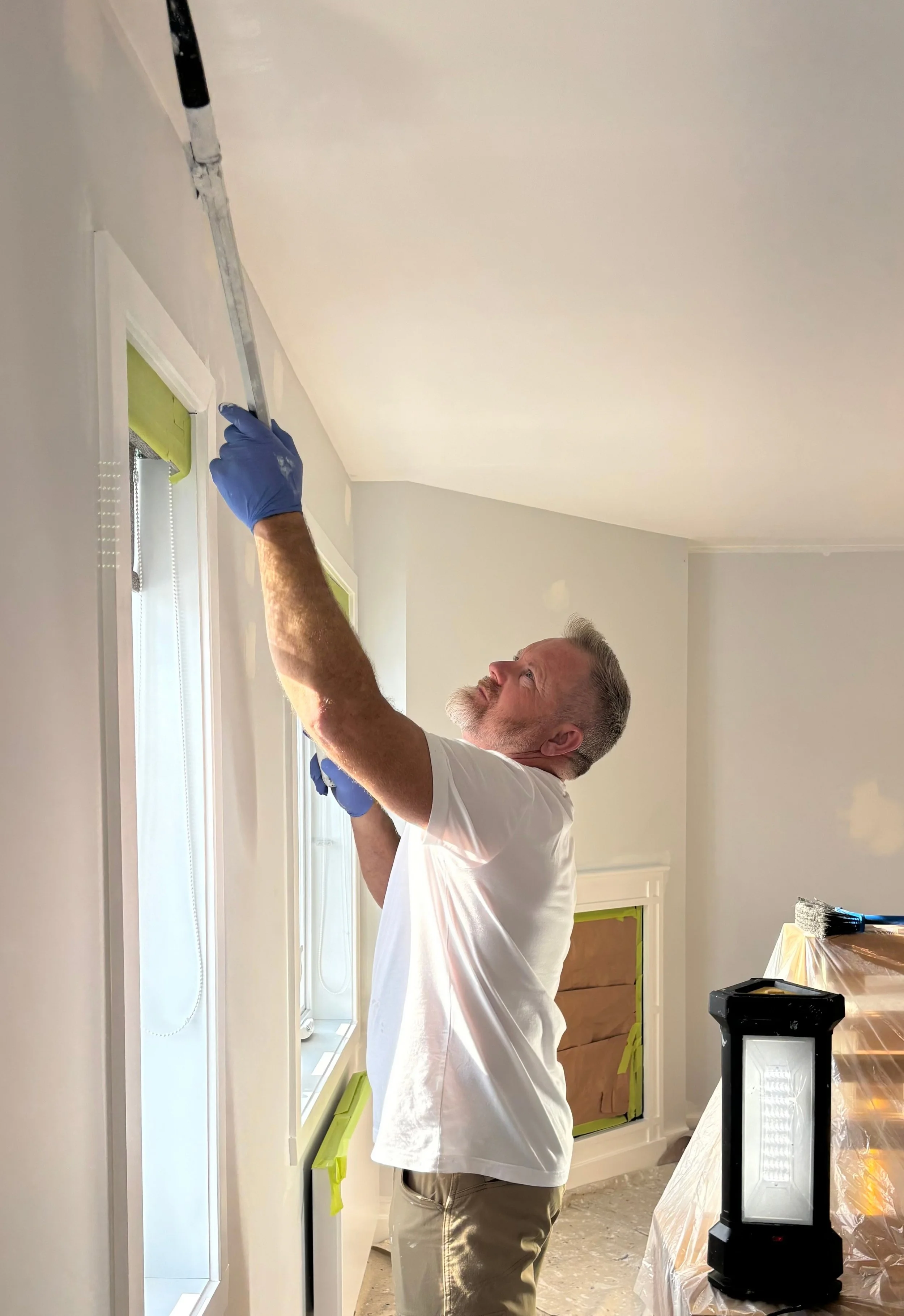 Patrick Jacob de MP Entretien Peinture appliquant la peinture au rouleau sur un mur près des fenêtres, cadrages et surfaces masqués au ruban vert