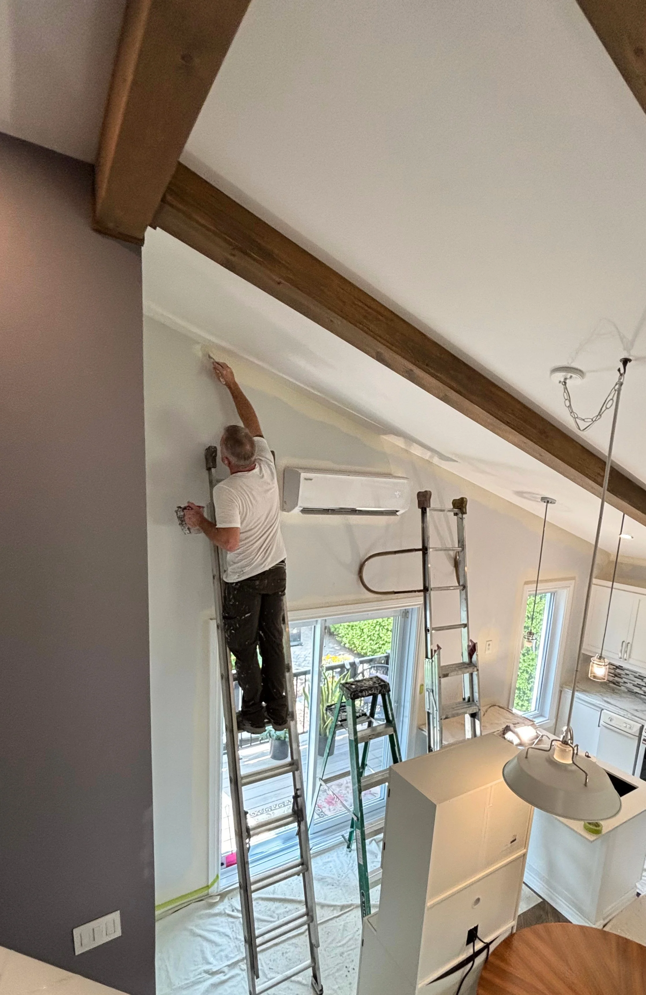 Patrick Jacob, copropriétaire de MP Entretien Peinture, découpant au pinceau le haut d'un mur sous un plafond cathédrale à poutres de bois dans une maison à Terrebonne