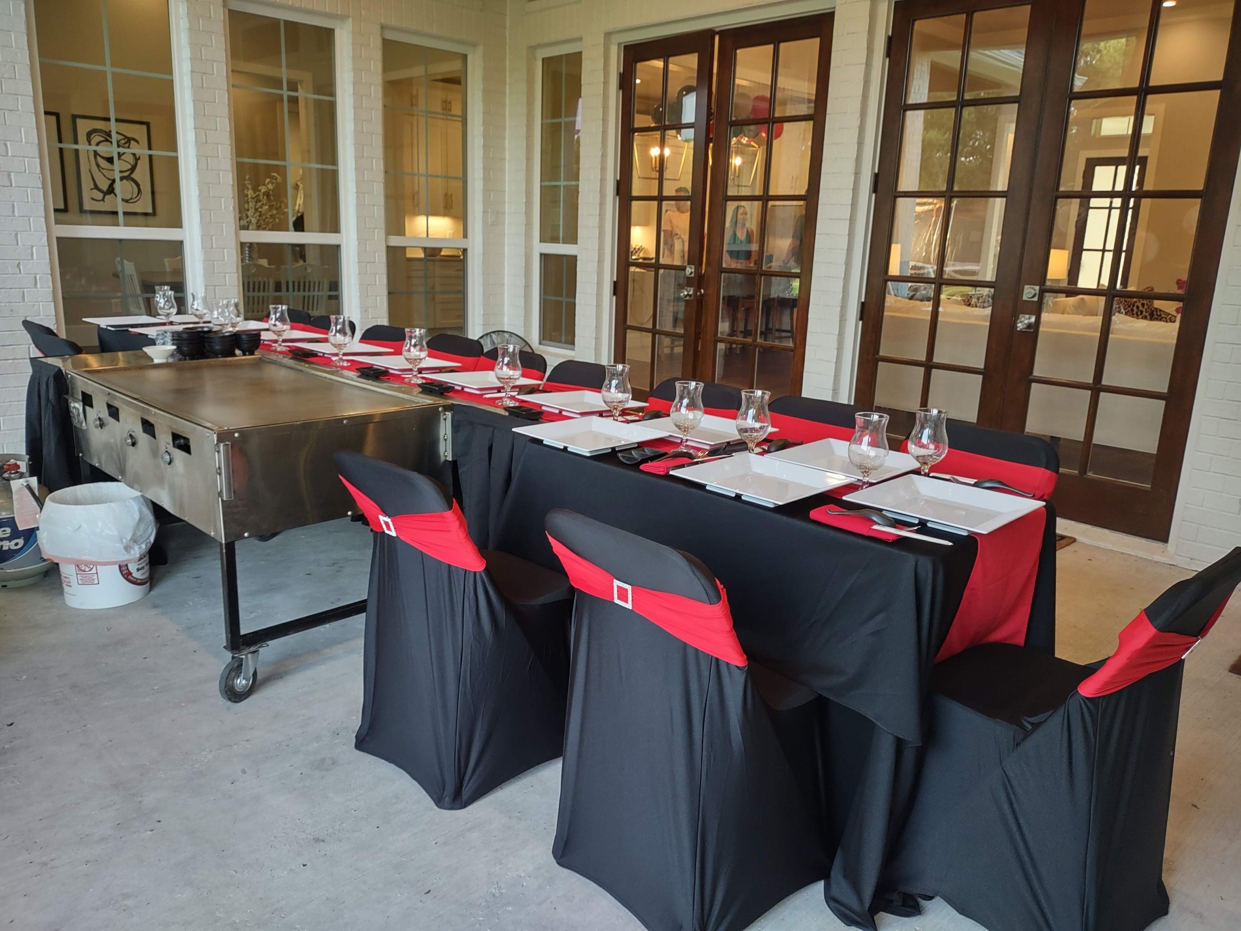 A dining table set for a meal, with black and red chair covers, white square plates, wine glasses, and red napkins, located in a bright room with white brick walls and large glass doors.