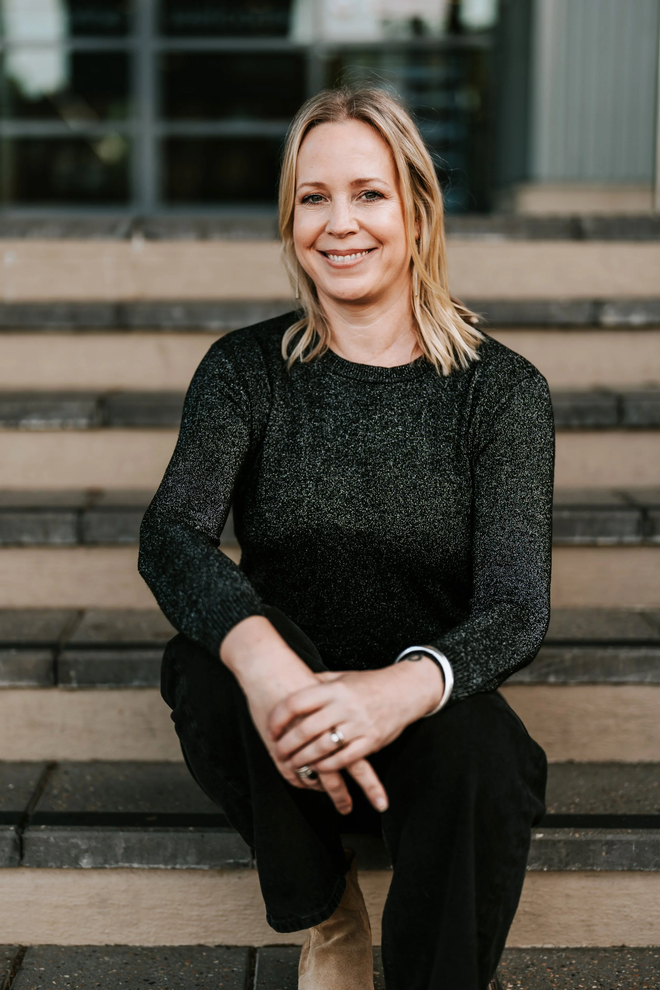 A woman with blonde hair, wearing a black sparkly long-sleeve top, sitting on outdoor steps, smiling at the camera.