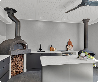 Modern kitchen with a wood-fired pizza oven, black chimney pipe, white cabinets, black countertops, and a kitchen island with a book and plants.