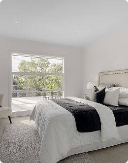 A bedroom with a large window, a bed with white bedding and black pillows, a nightstand with a lamp, and a white chair near the window.