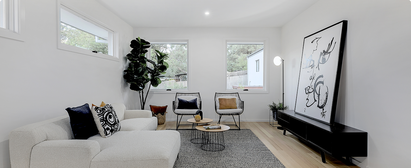 Modern minimalist living room with white walls, light wood flooring, a white sofa with black and patterned pillows, two black wire chairs with cushions, a large black TV stand with artistic framed artwork on top, a tall floor lamp, a small round coffee table, and large windows with trees outside.
