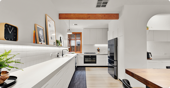 Modern kitchen with white cabinets, black appliances, and wooden accents, featuring a long countertop, wall clock, and framed art.