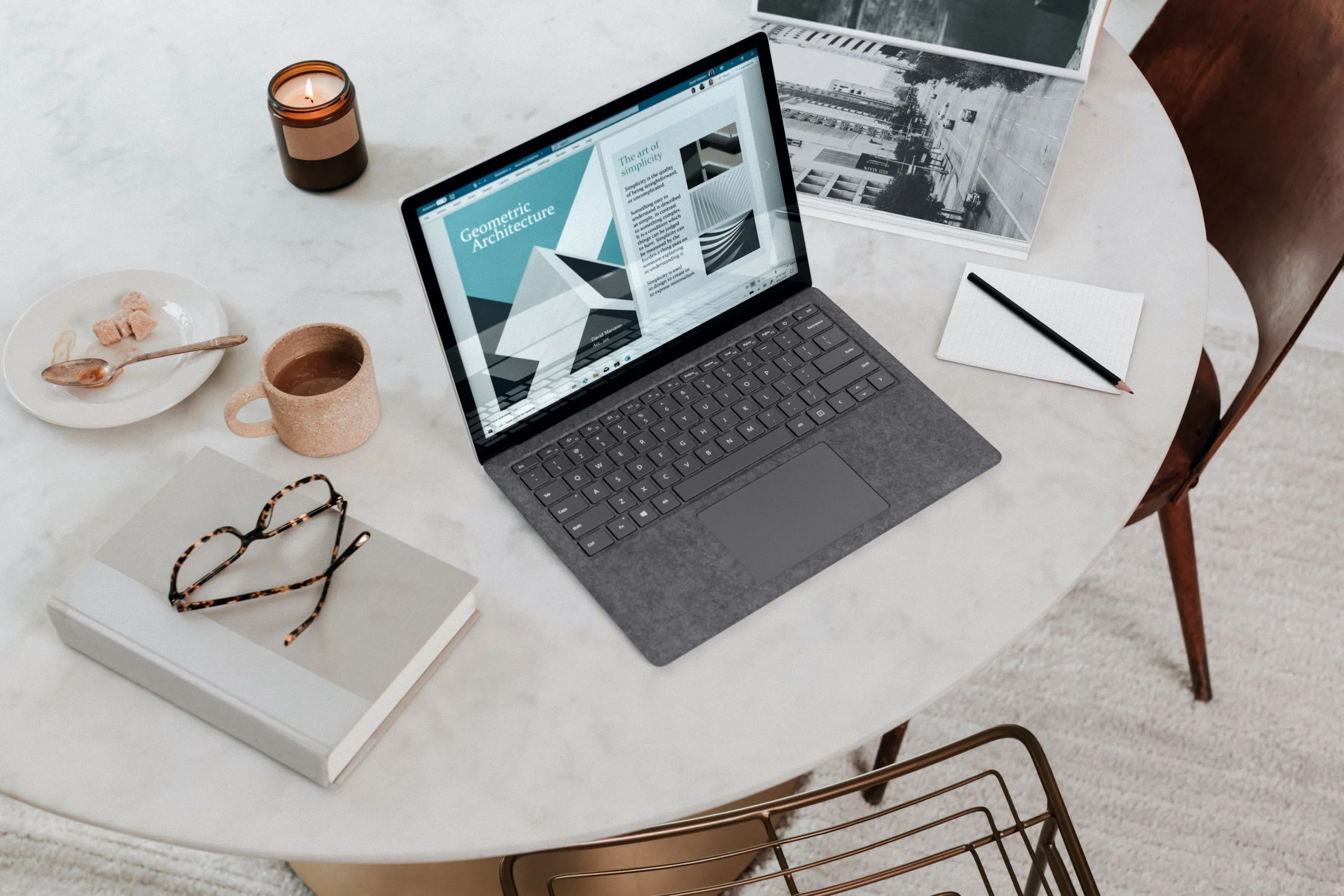 A white round table with a laptop displaying a webpage about geometric architecture, a candle, a ceramic mug with a beverage, a small plate with some snack pieces and a spoon, a white book with tortoise-shell glasses on top, a notepad with a black pencil, and some black and white photographs of urban streets are arranged on it.
