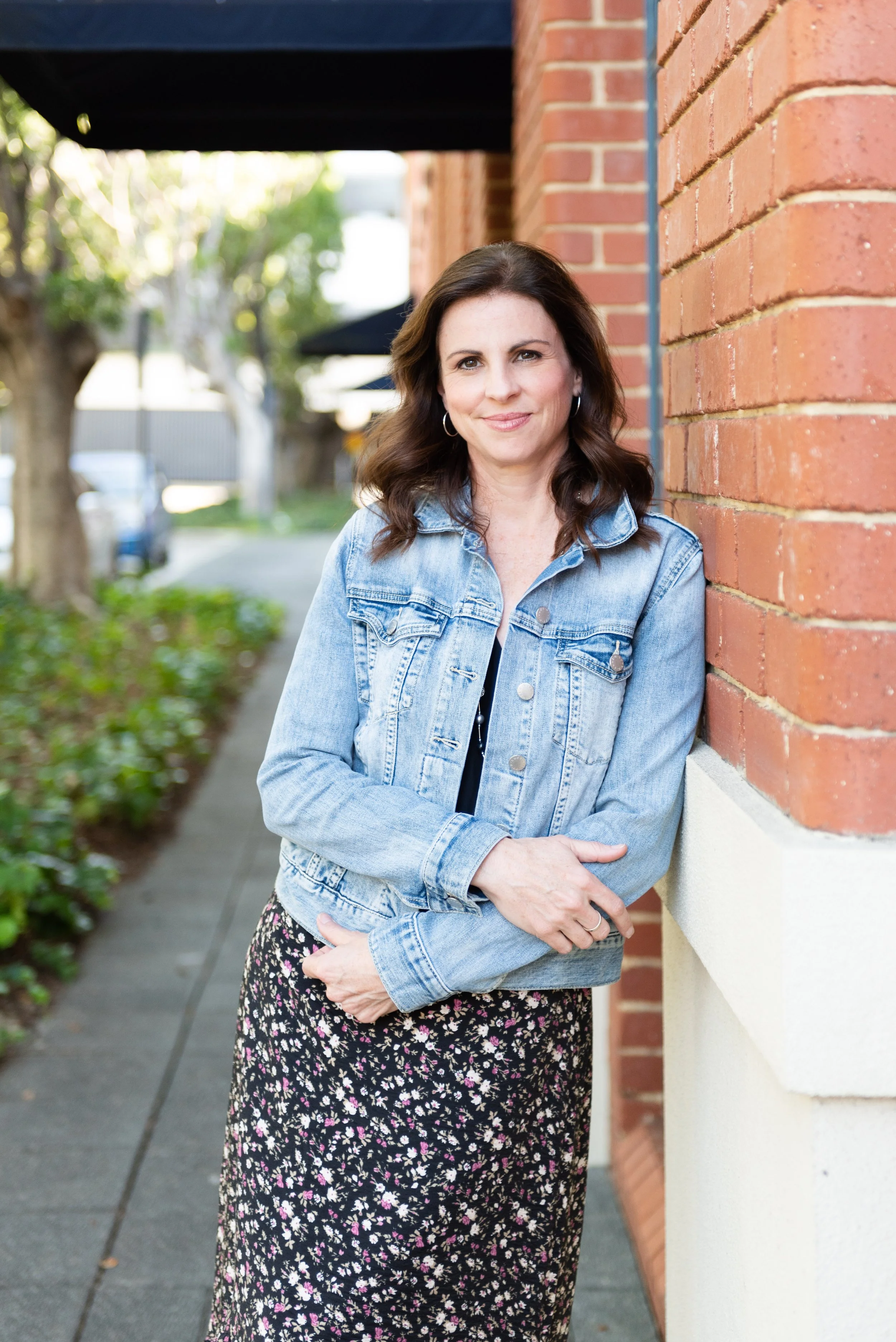 A woman with dark brown hair, wearing a denim jacket and a floral skirt, leaning against a brick wall outdoors, with trees and parked cars in the background.