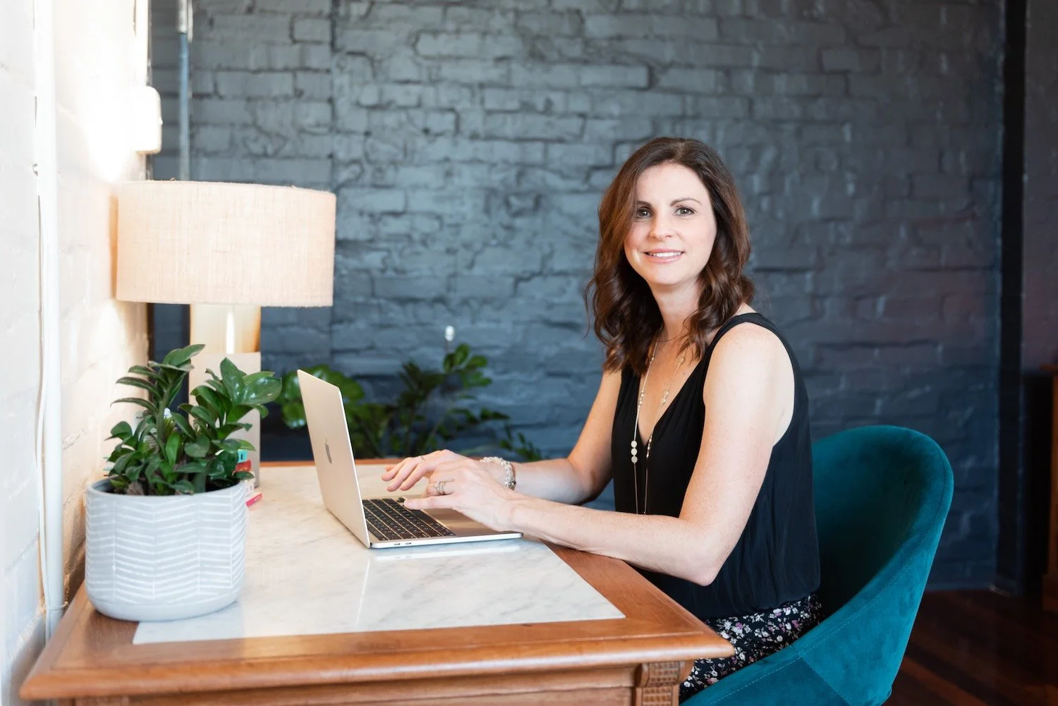 A woman with brown hair wearing a black sleeveless top sitting at a wooden desk using a silver laptop. The desk has a white plant in a striped pot and a table lamp with a beige shade. She is smiling and looking at the camera, with a dark gray brick wall in the background and some green plants behind her.