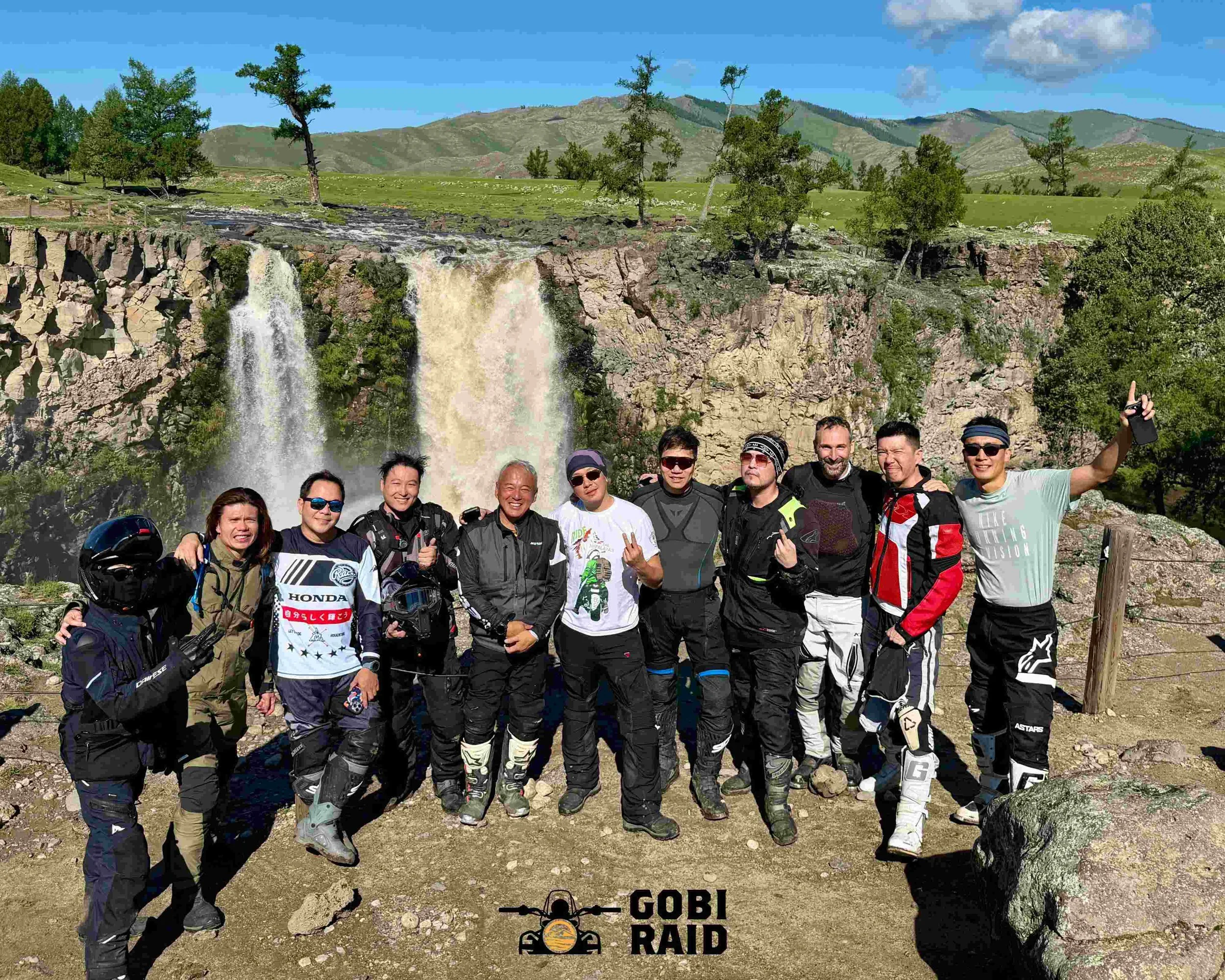 A group of motorcycle riders posing in front of a waterfall with mountains and trees in the background.