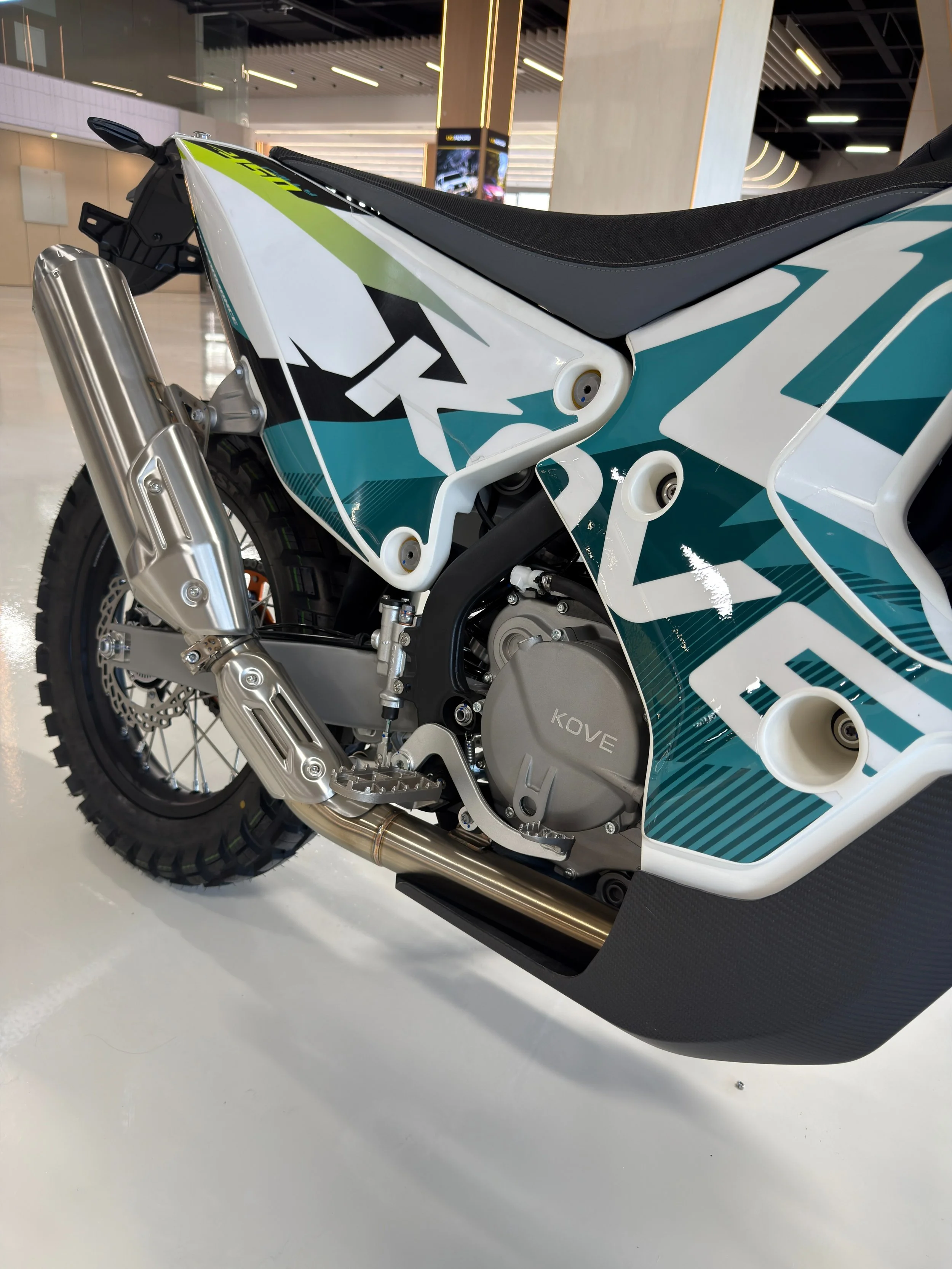 Close-up of a rally bike showing part of the rear wheel, exhaust, and side panel with graphics. KOVE Rally 450 RE