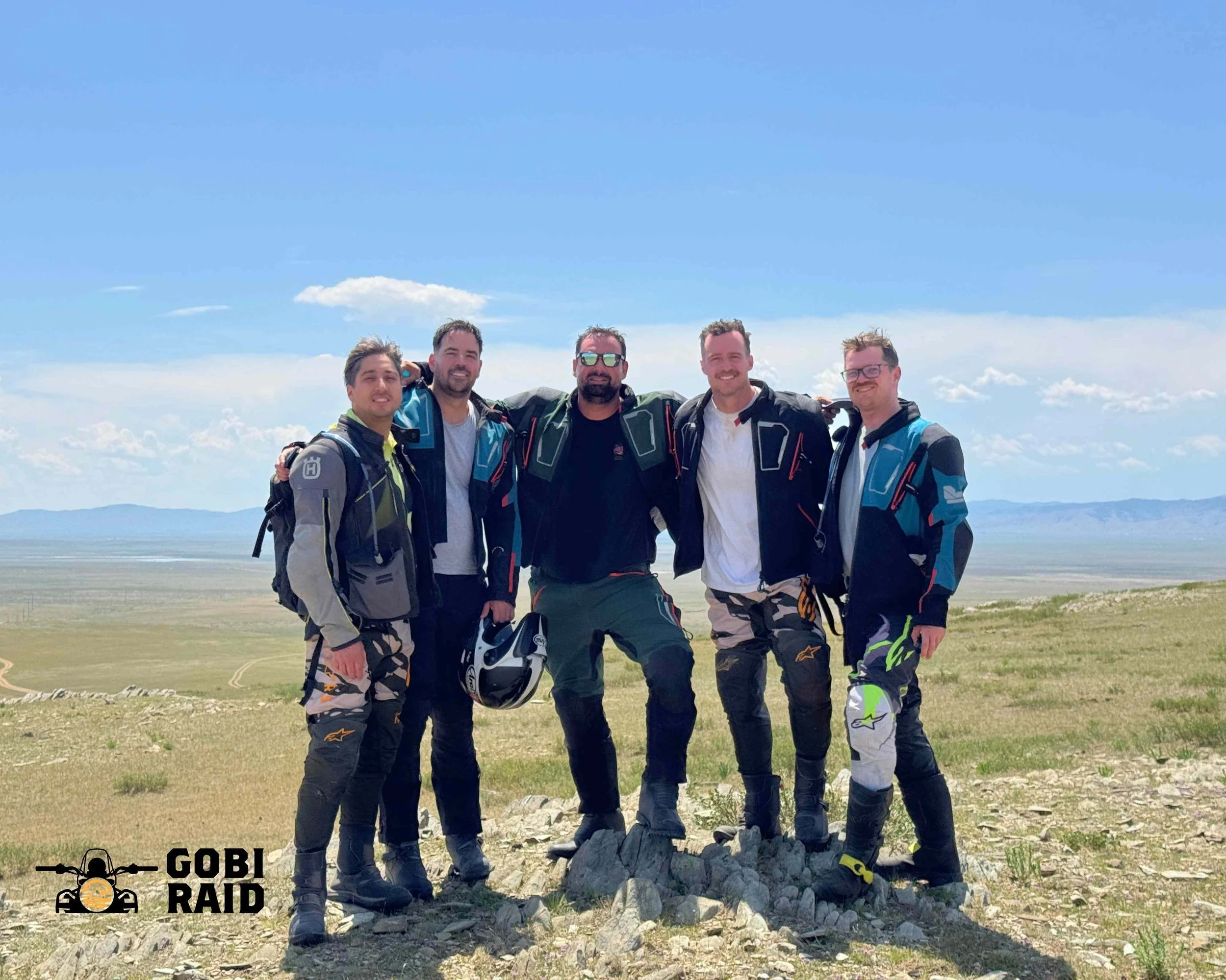 Five men standing outdoors on a grassy landscape with a clear blue sky behind them, dressed in motorcycle riding gear and smiling at the camera, with some holding their motorcycle helmets.