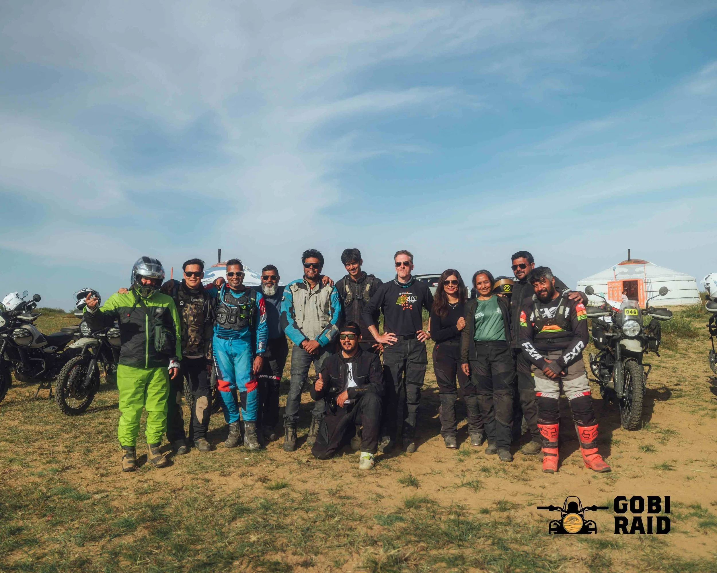 A group of twelve motorcyclists in riding gear posing together outdoors. There are motorcycles parked on a grassy field behind them, with a couple of small tents or structures. The sky is mostly cloudy with patches of blue. A logo reading 'Gobi Rally