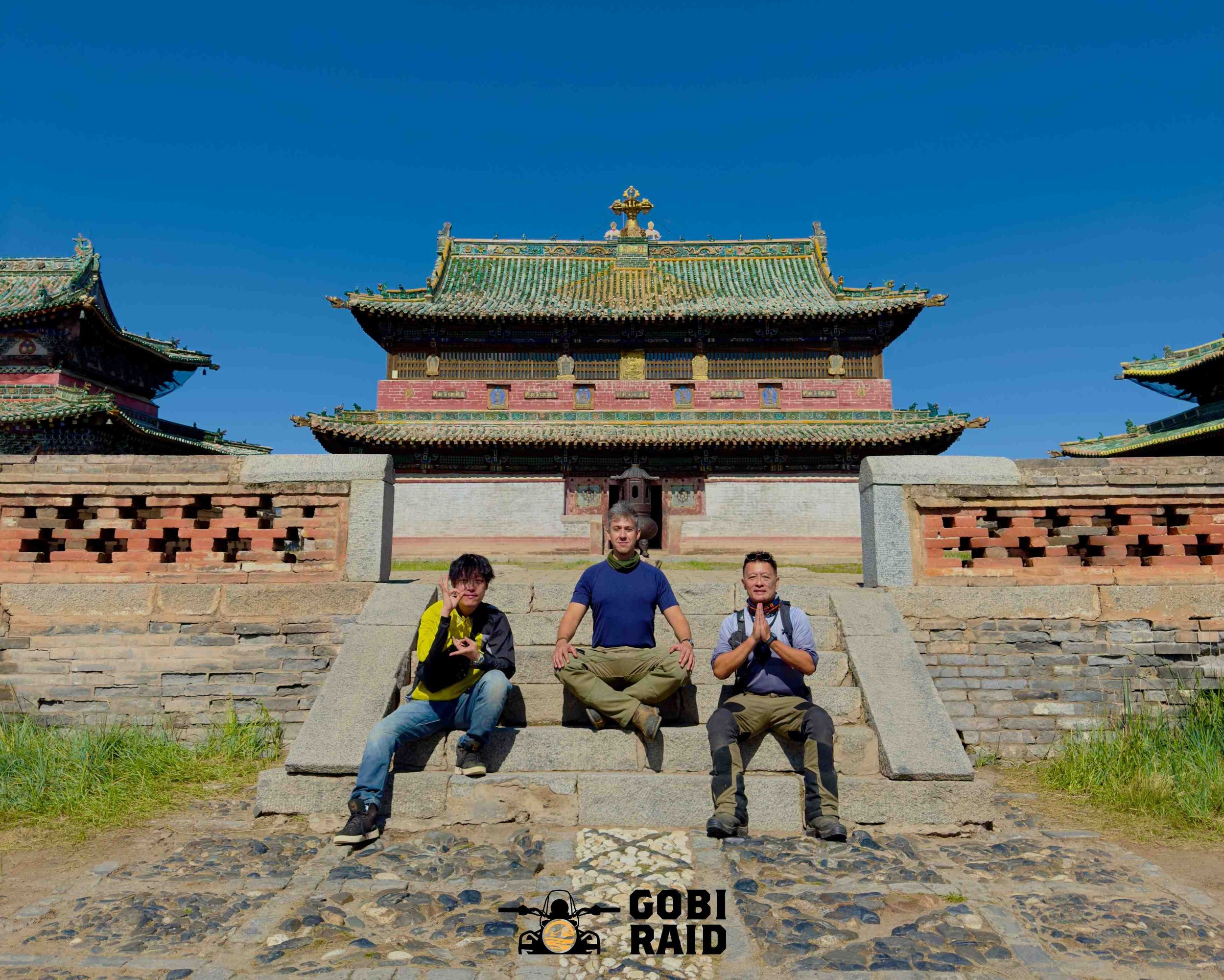 Three people sitting on stone steps in front of a traditional Asian temple with ornate, colorful roof details and a blue sky overhead.