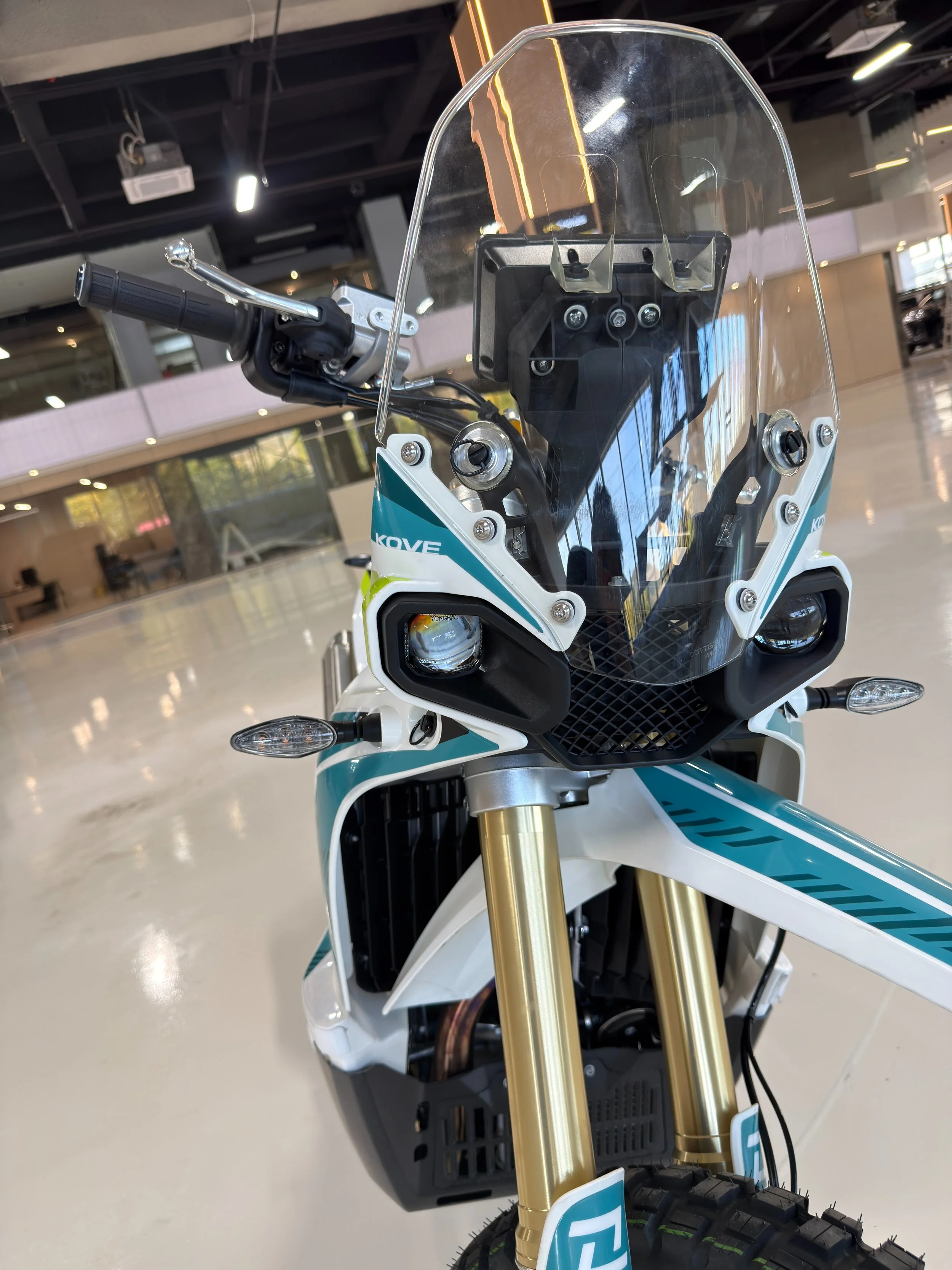 Front view of an rally off-road motorcycle with a transparent windshield, gold forks, and a black and white body, displayed indoors. KOVE Rally 450 RE