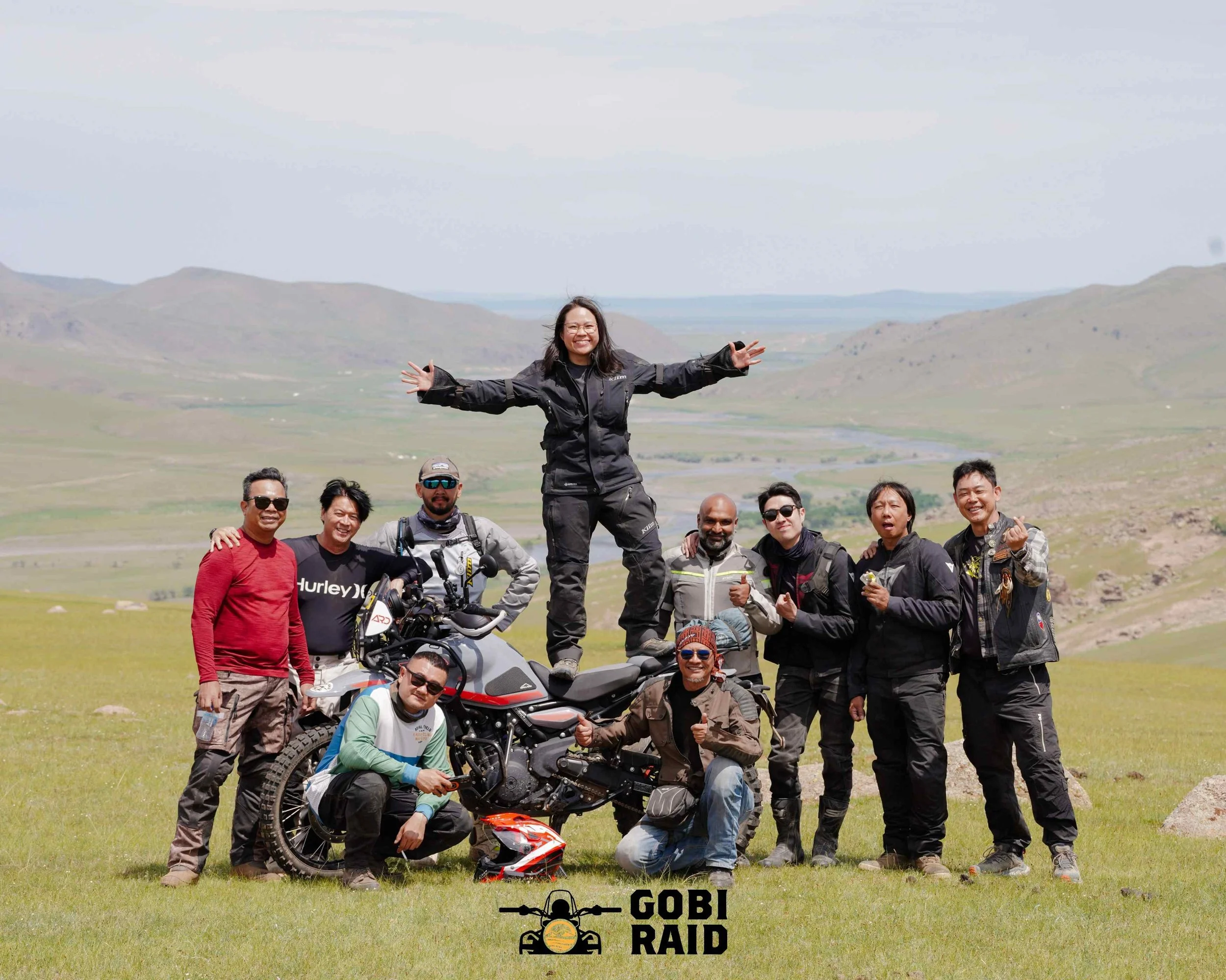 Group of people outdoors in green fields with mountains in the background, some posing and smiling, a woman standing with arms outstretched, motorcycle and helmets in the foreground, and a logo that says 'Gobi Road Raid' at the bottom.