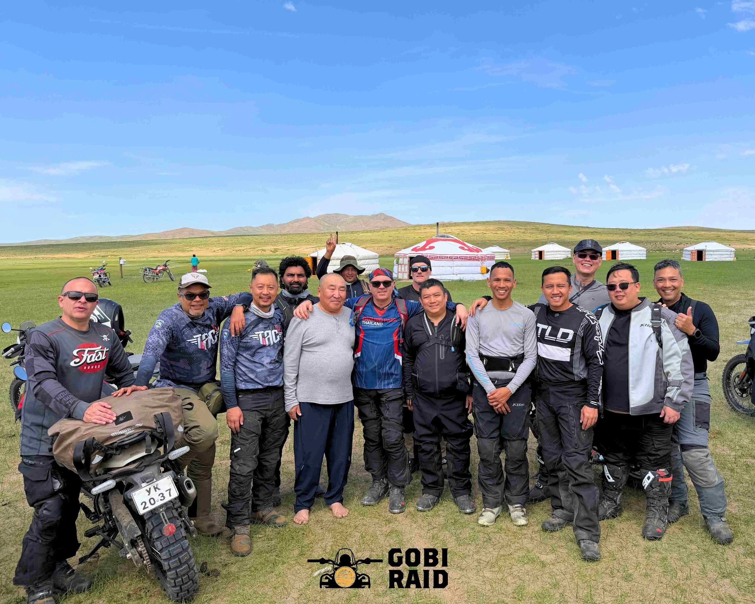 Group of men standing outdoors in a grassy field with yurts, motorcycles, and mountains in the background under a blue sky. Logo at the bottom reads "Gobi Ultra Raid."