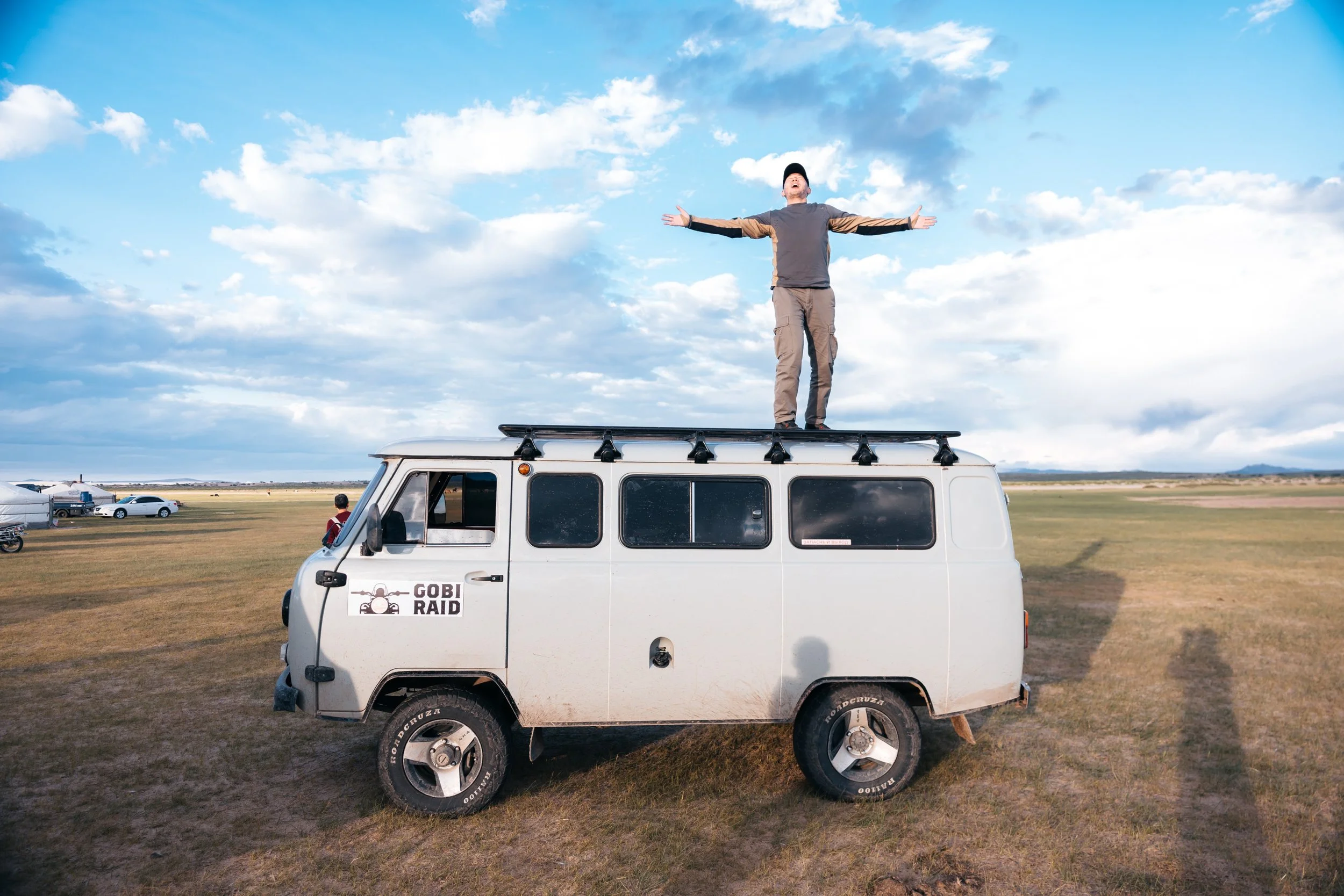 A person standing on top of a Russian UAZ vehicle with arms outstretched, outdoors in an open field under a partly cloudy sky during daytime.