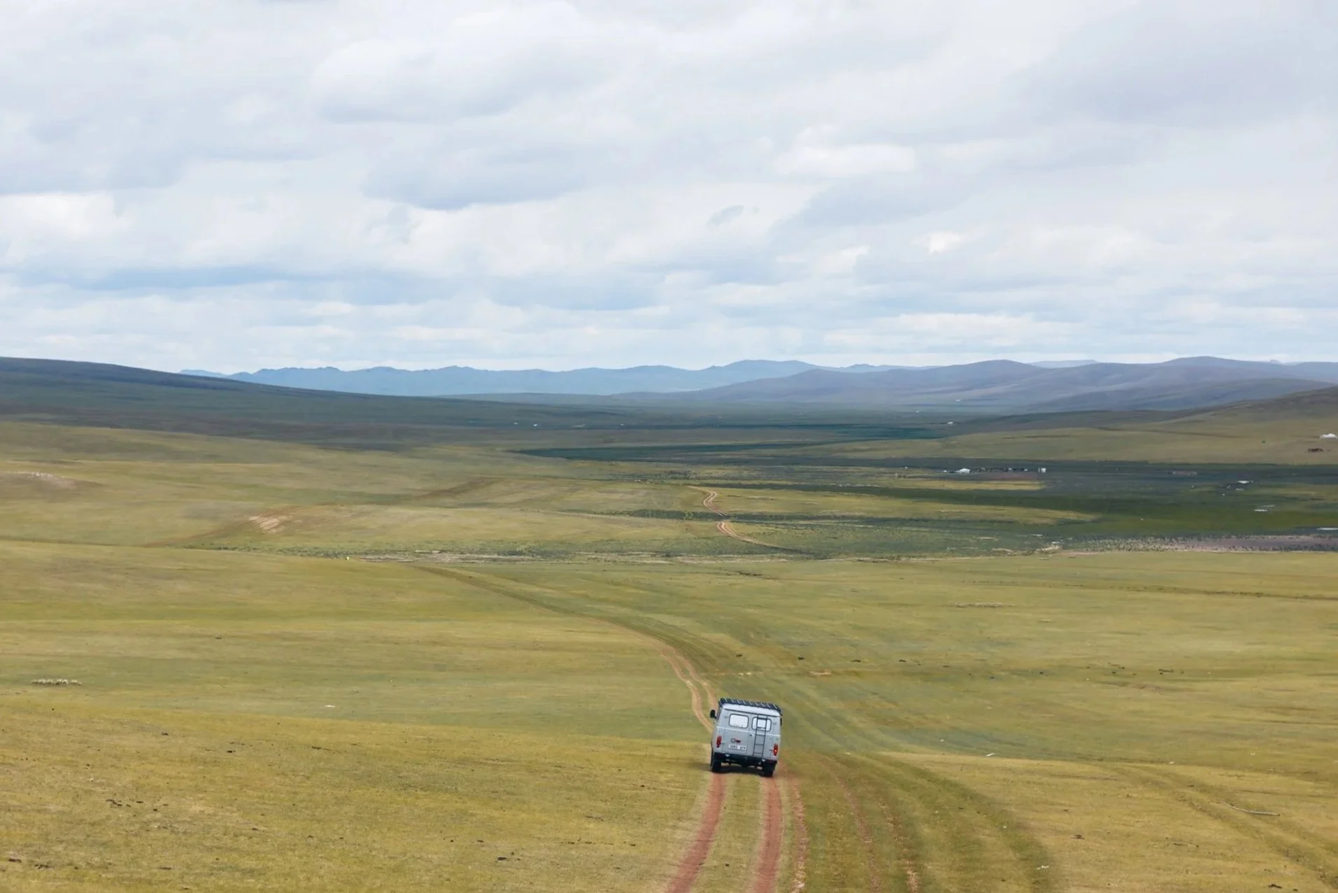 Russian UAZ driving on a dirt road through a vast open grassy landscape with rolling hills and mountains in the distance under a cloudy sky.