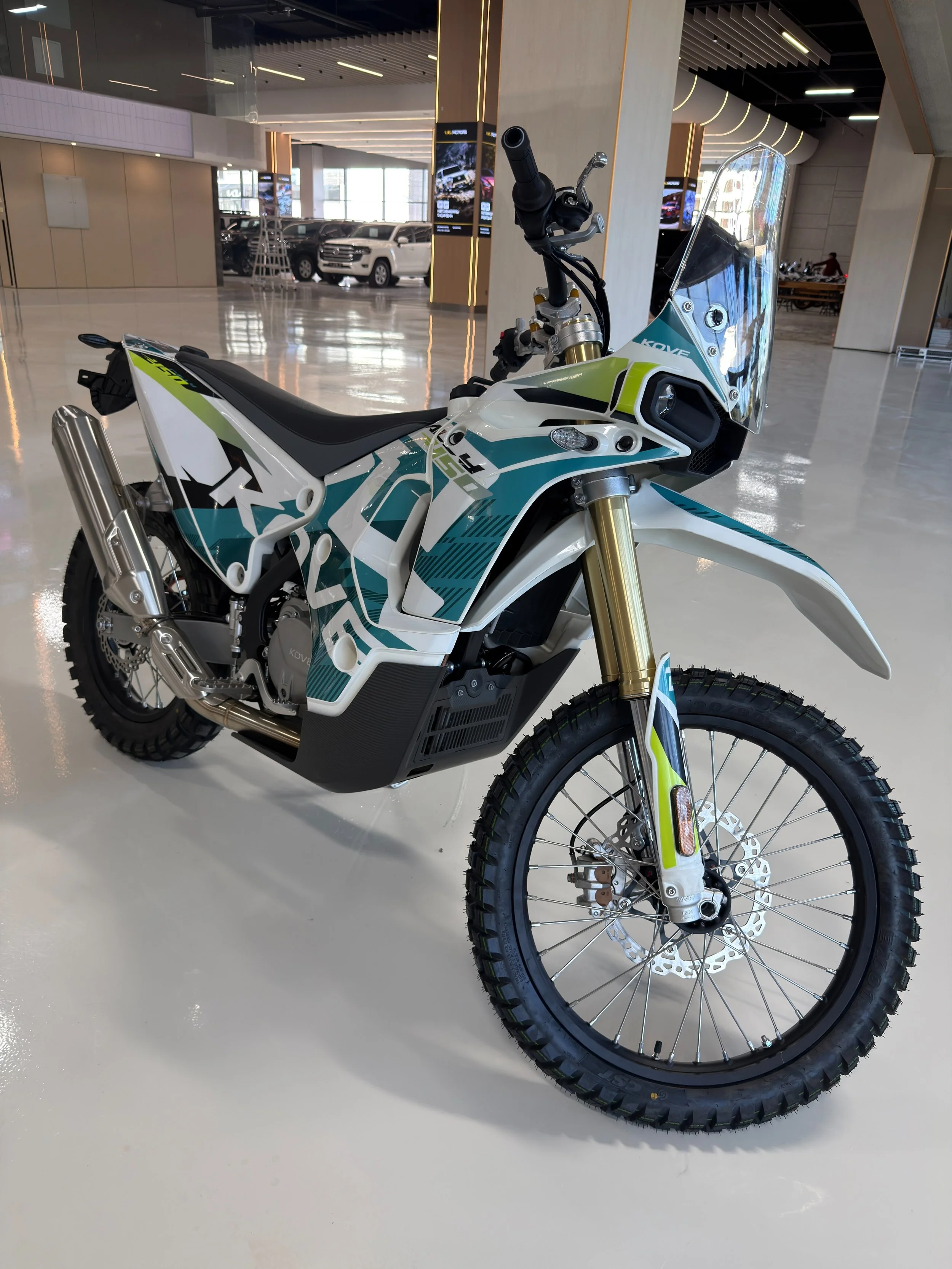 Modern off-road motorcycle with a white and teal body, gold suspension forks, knobby tires, and a clear windshield, displayed indoors at a showroom. KOVE Rally 450 RE