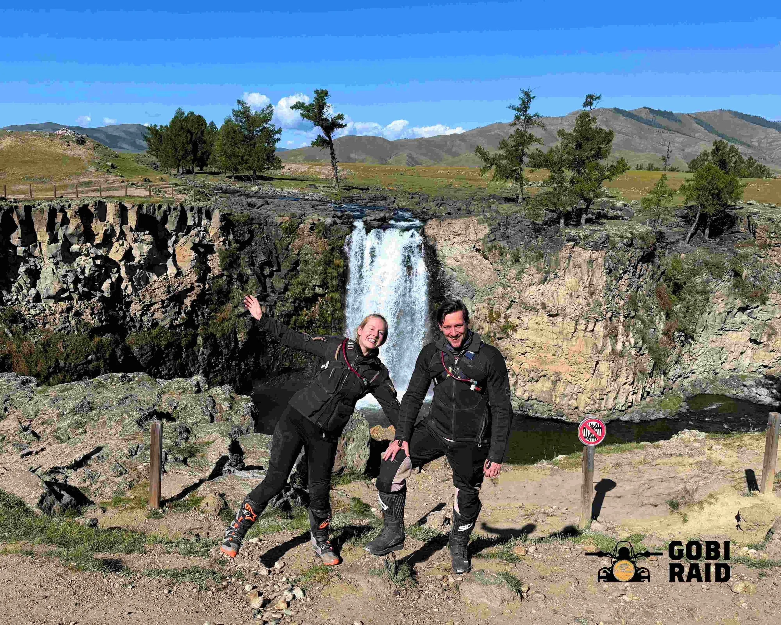 Two smiling people in motorcycle gear posing near a waterfall in a mountainous landscape during daytime.