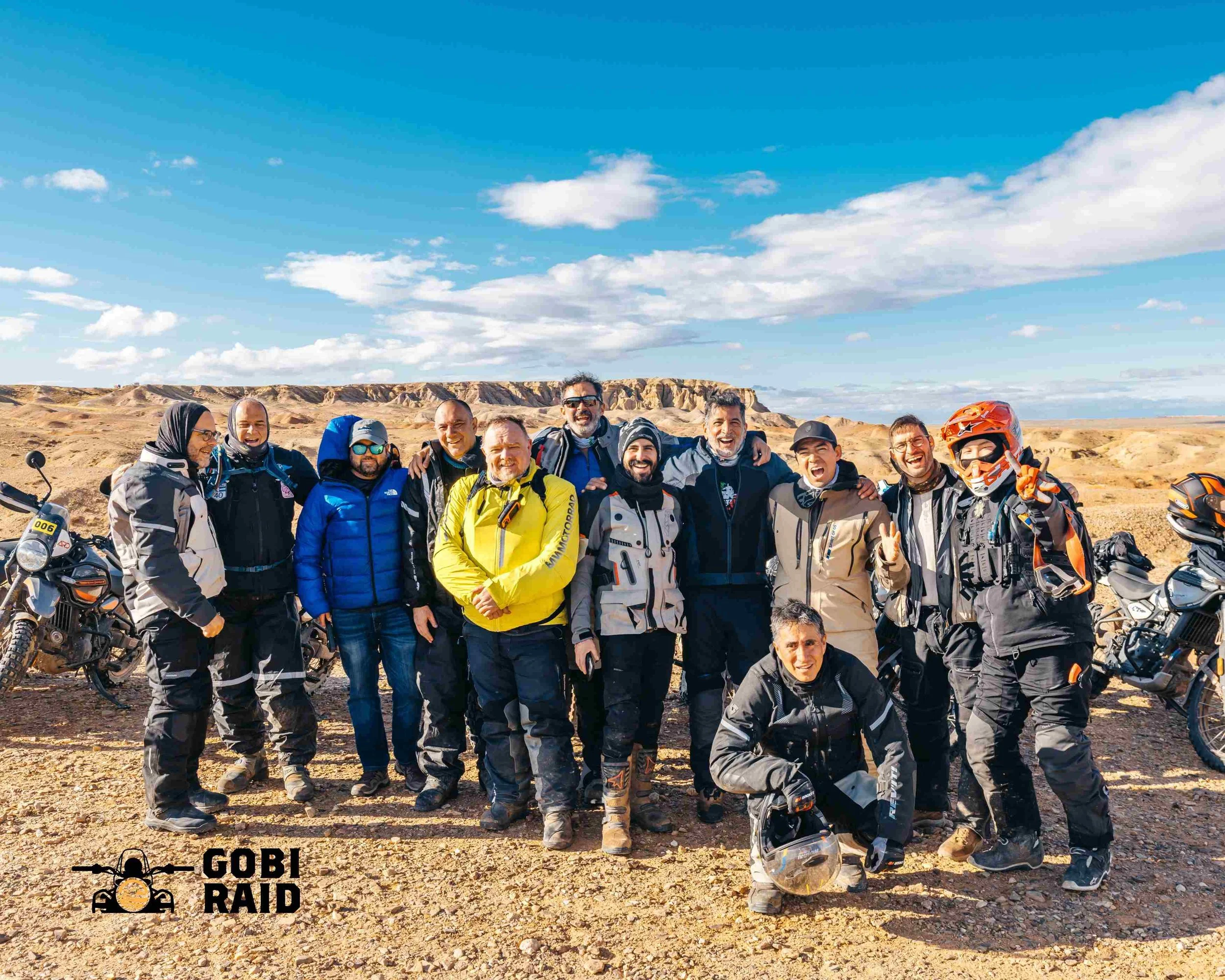 Group of people in outdoor landscape with rocky hills and blue sky, standing with motorcycles, some wearing riding gear and helmets, smiling and posing for the photo.