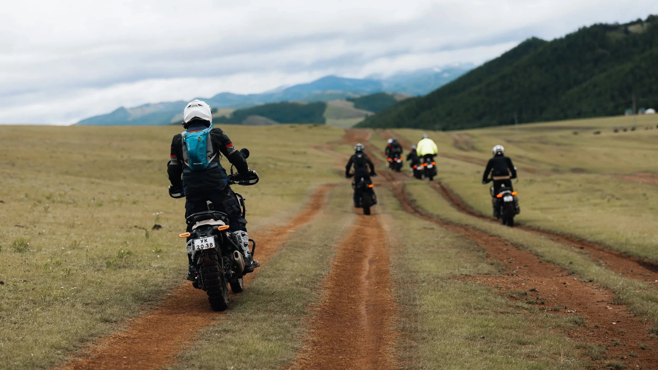 A group of motorcyclists riding on a dirt trail through a grassy field, with mountains and cloudy sky in the background.
