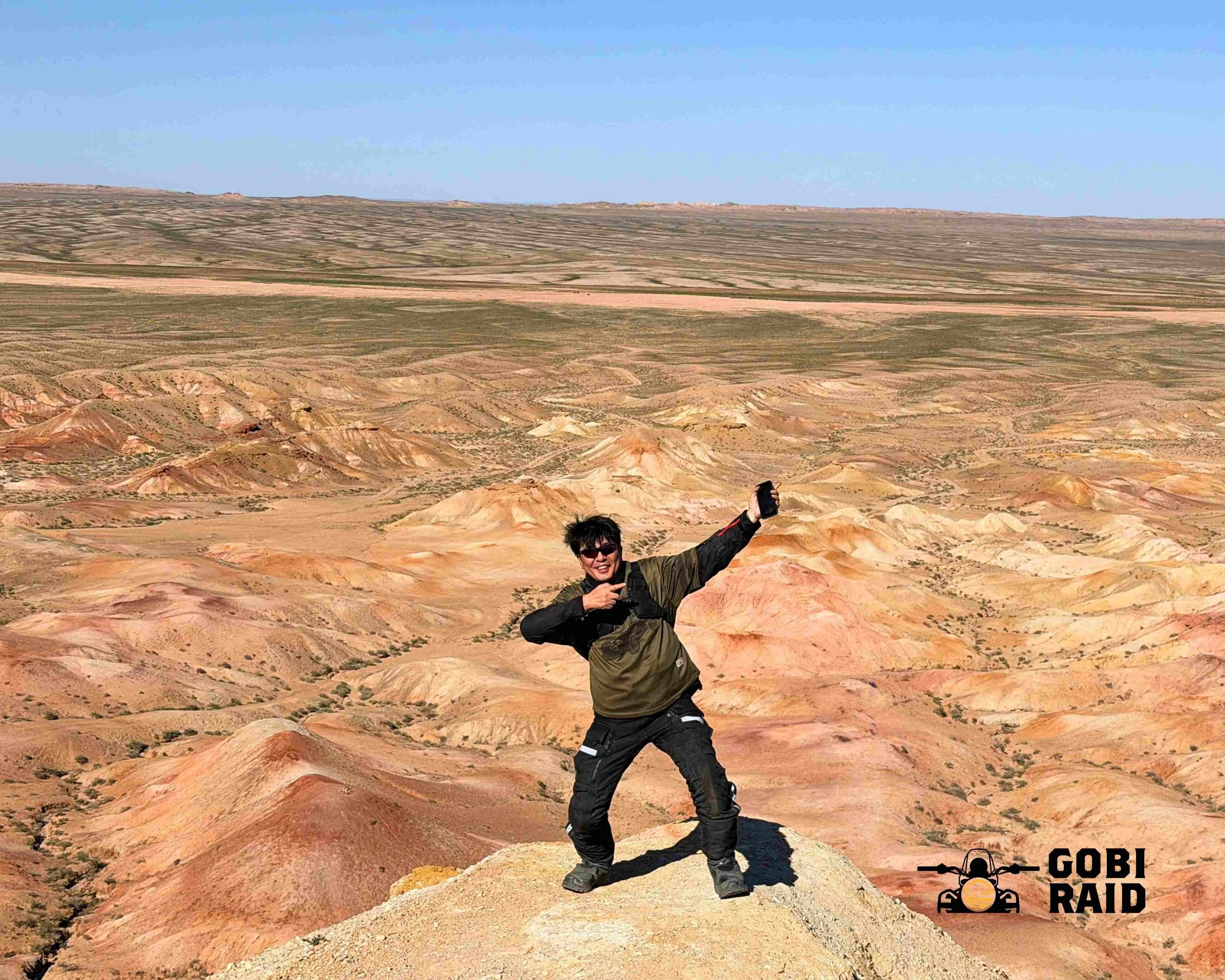 Person wearing sunglasses and a jacket taking a selfie or photo in a desert landscape with colorful layered rock formations and a blue sky in the background.
