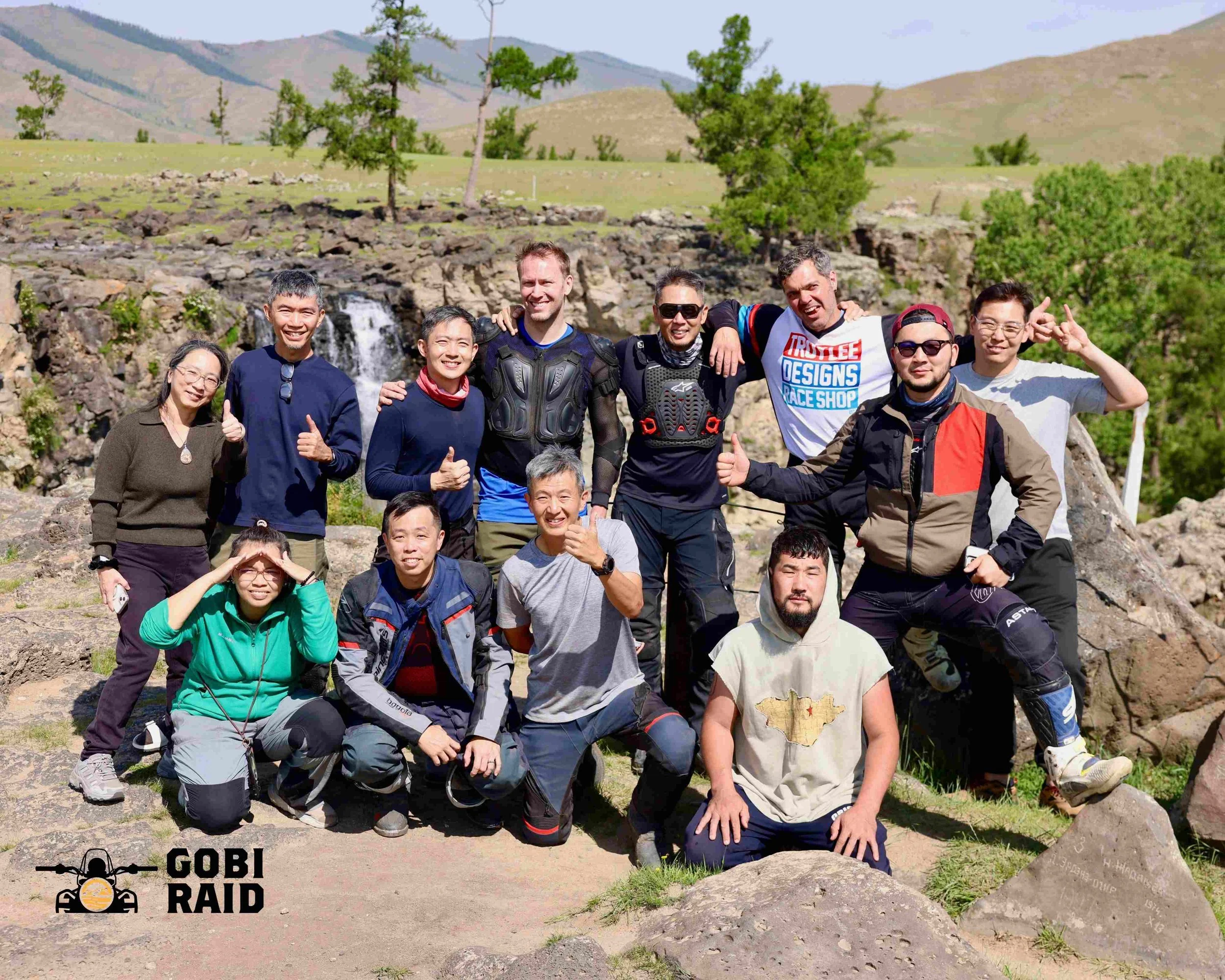 A group of people outdoors on a sunny day, posing for a photo with a mountain landscape and trees in the background, some making thumbs-up gestures, others smiling or making various hand signs, with a logo in the bottom left corner that says "Gobi Ra
