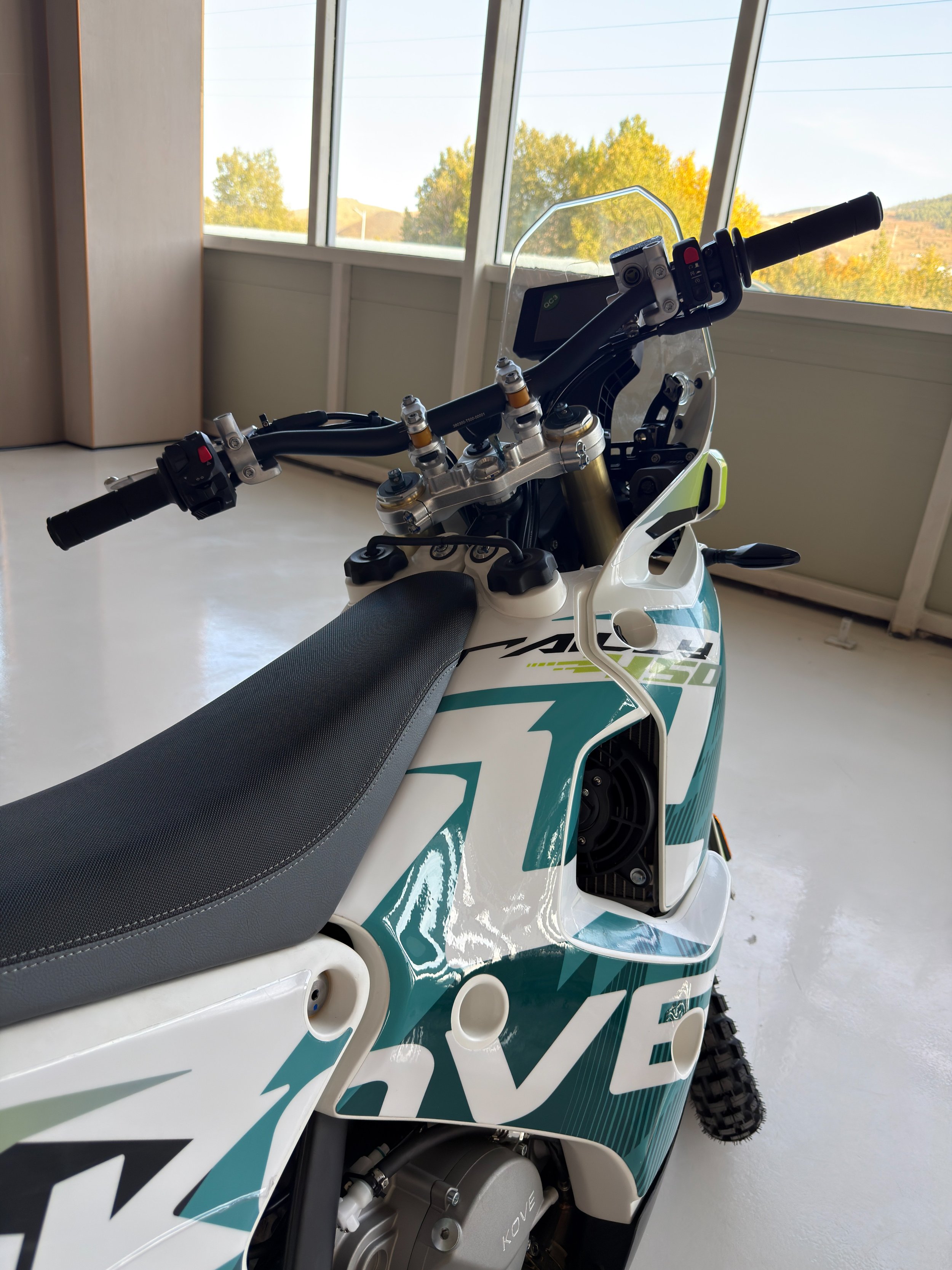 Close-up of a rally bike's handlebar and front section, showing a digital display, engine components, and body graphics with the logo 'KOVE'. A shielding screen is attached to the handlebar. KOVE Rally 450 RE