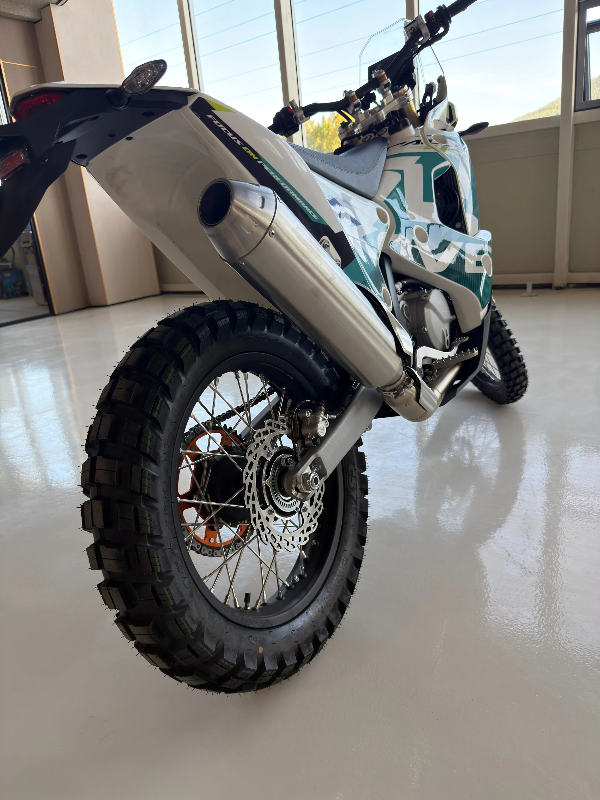Close-up of a rally motorcycle with large knobby tires, a white body with black and teal graphics, a large front brake disc, and an exhaust pipe. The bike is indoors near large windows. KOVE Rally 450 RE