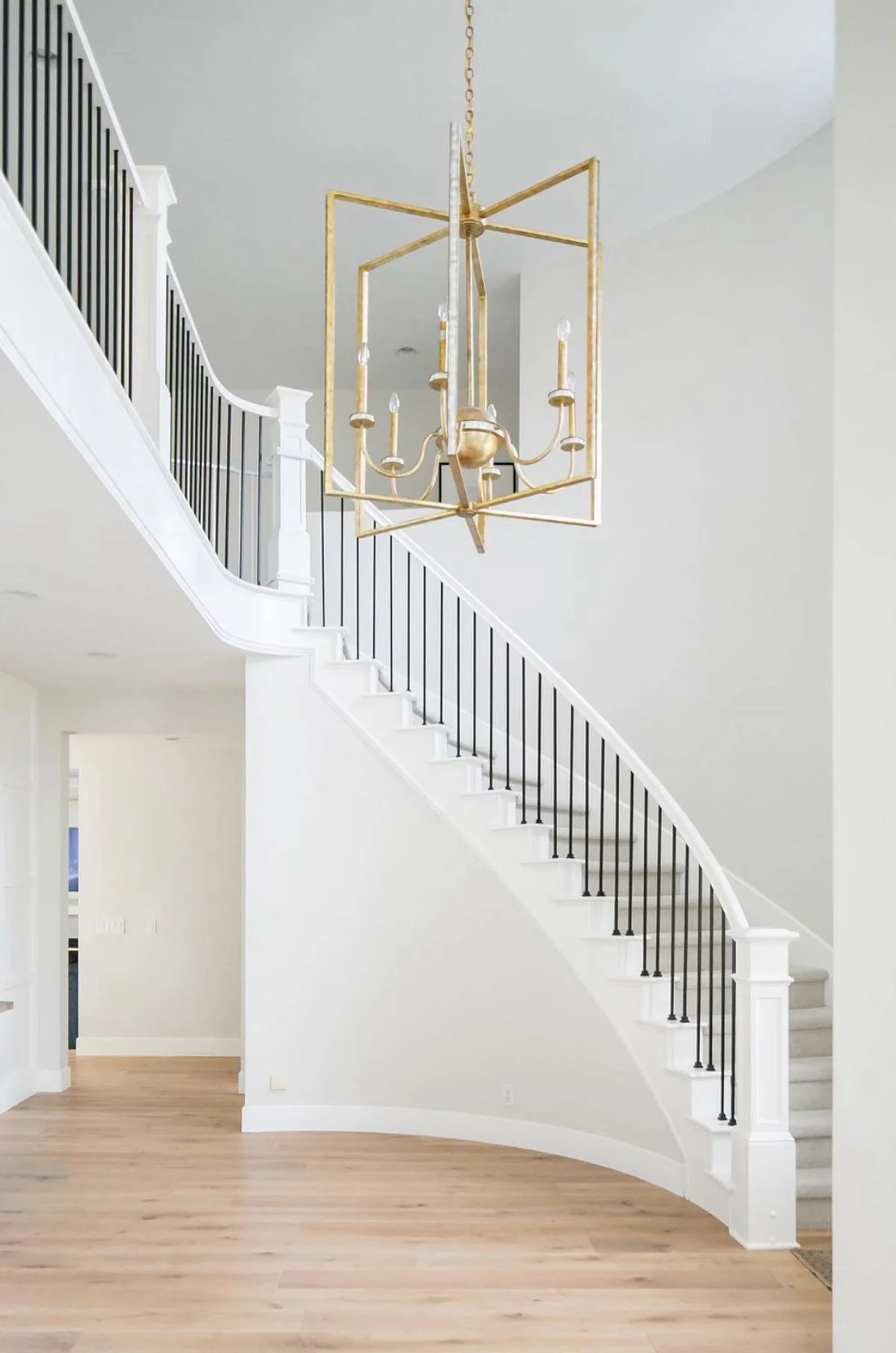 Interior view of a modern home with a curved staircase, white walls, a wooden floor, and a gold chandelier hanging from the ceiling.