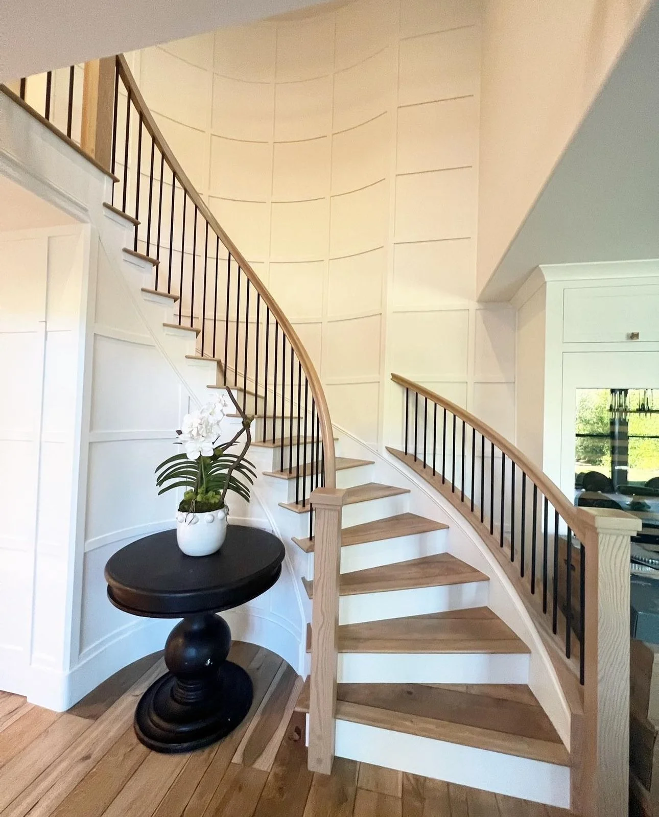 Interior view of a staircase with wooden steps and black metal railing, beside a white wall and a window reflecting greenery outside, with a small black table holding a white potted orchid plant.