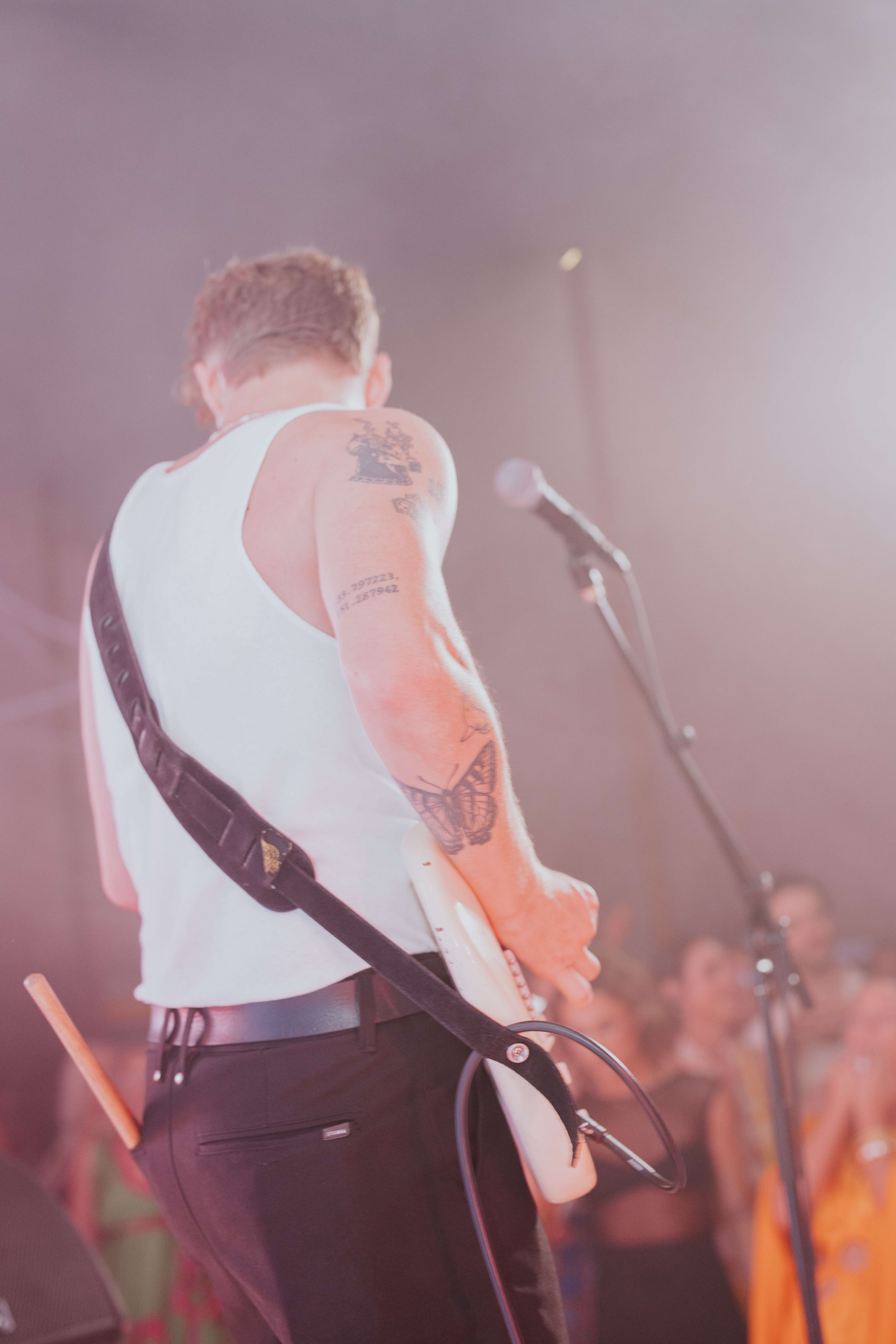 A person playing electric guitar on stage, wearing a white tank top and black pants, with visible tattoos on their arm, and a drumstick in their back pocket. Microphone and audience slightly blurred in the background.