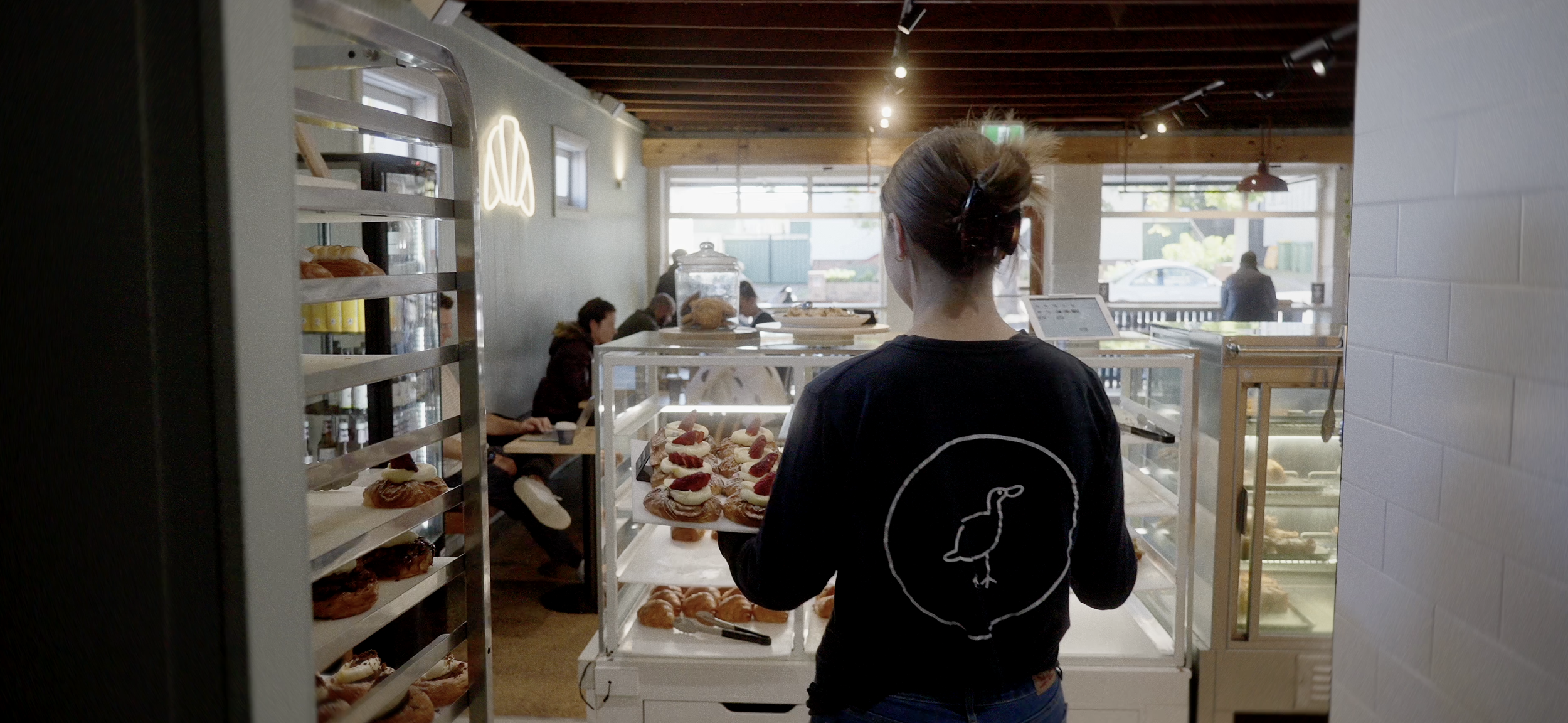 Person holding a tray of pastries in a bakery with display cases and customers seated in the background.