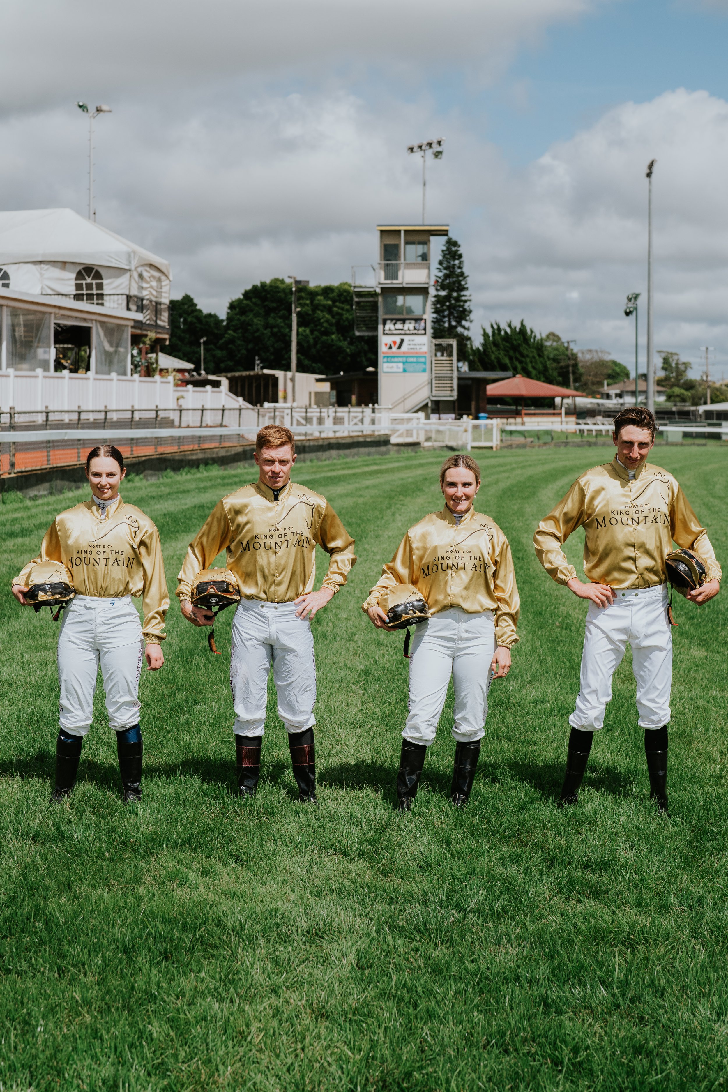Four jockeys wearing gold jackets and white pants standing on a grassy racetrack, holding helmets. A racetrack building is visible in the background.