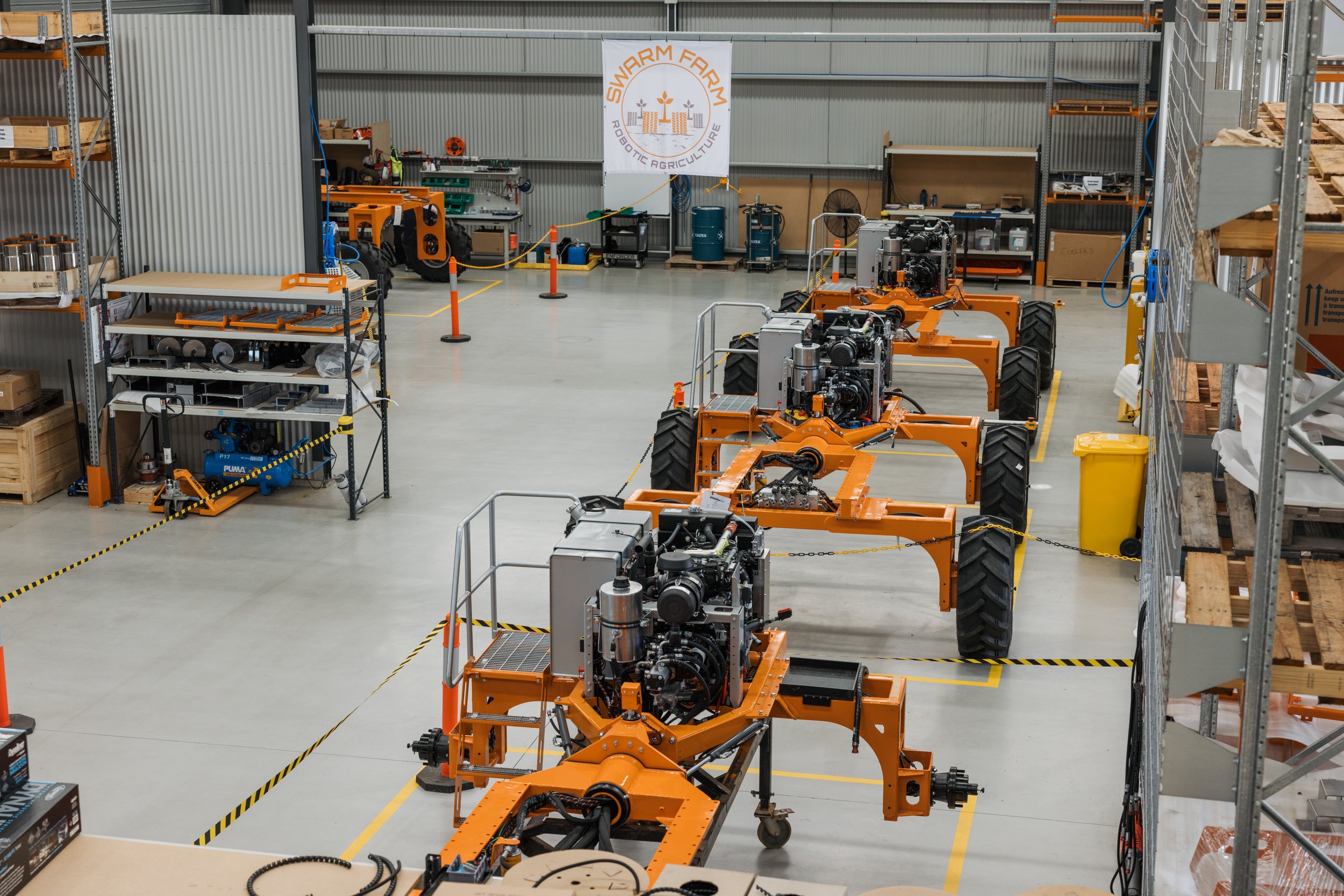 Interior of a warehouse featuring several orange robotic agricultural machines with large wheels, shelves with equipment, and a banner that reads 'Swarm Farm Robotic Agriculture.'