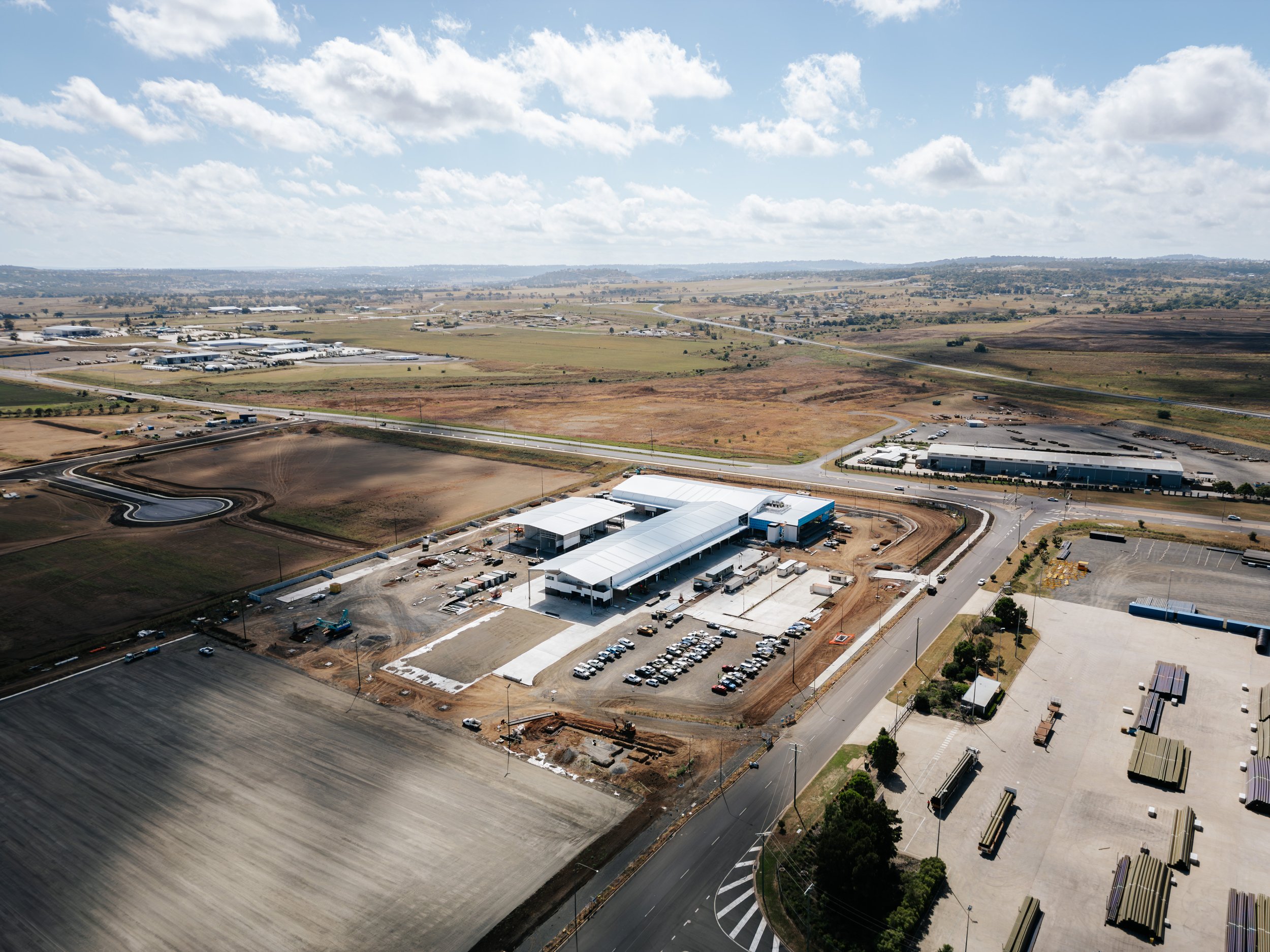 Aerial view of an industrial complex with buildings, roads, and parking areas surrounded by open fields under a cloudy sky.