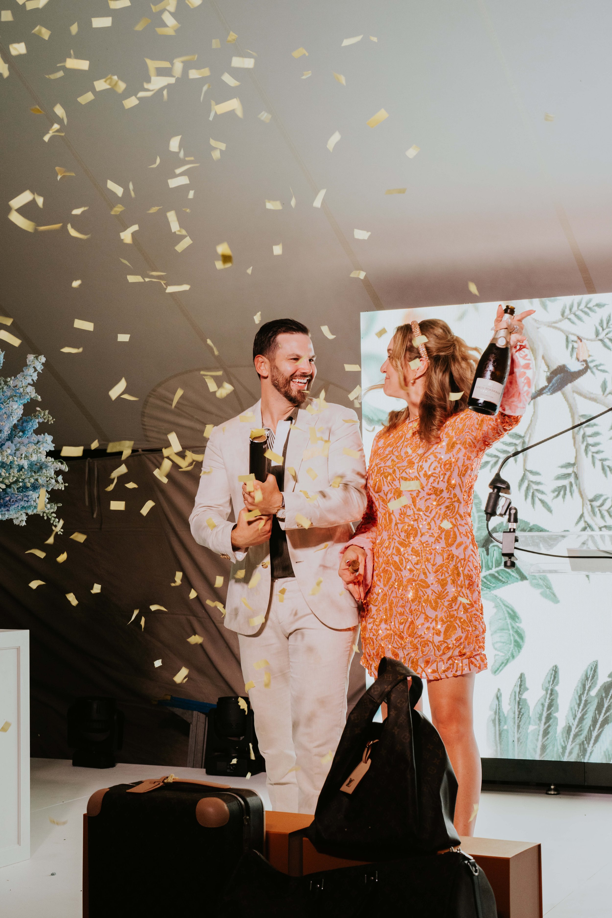 A man and a woman smiling and celebrating, holding a champagne bottle with confetti falling around them. They stand on a stage with decorative elements and luggage in the foreground.