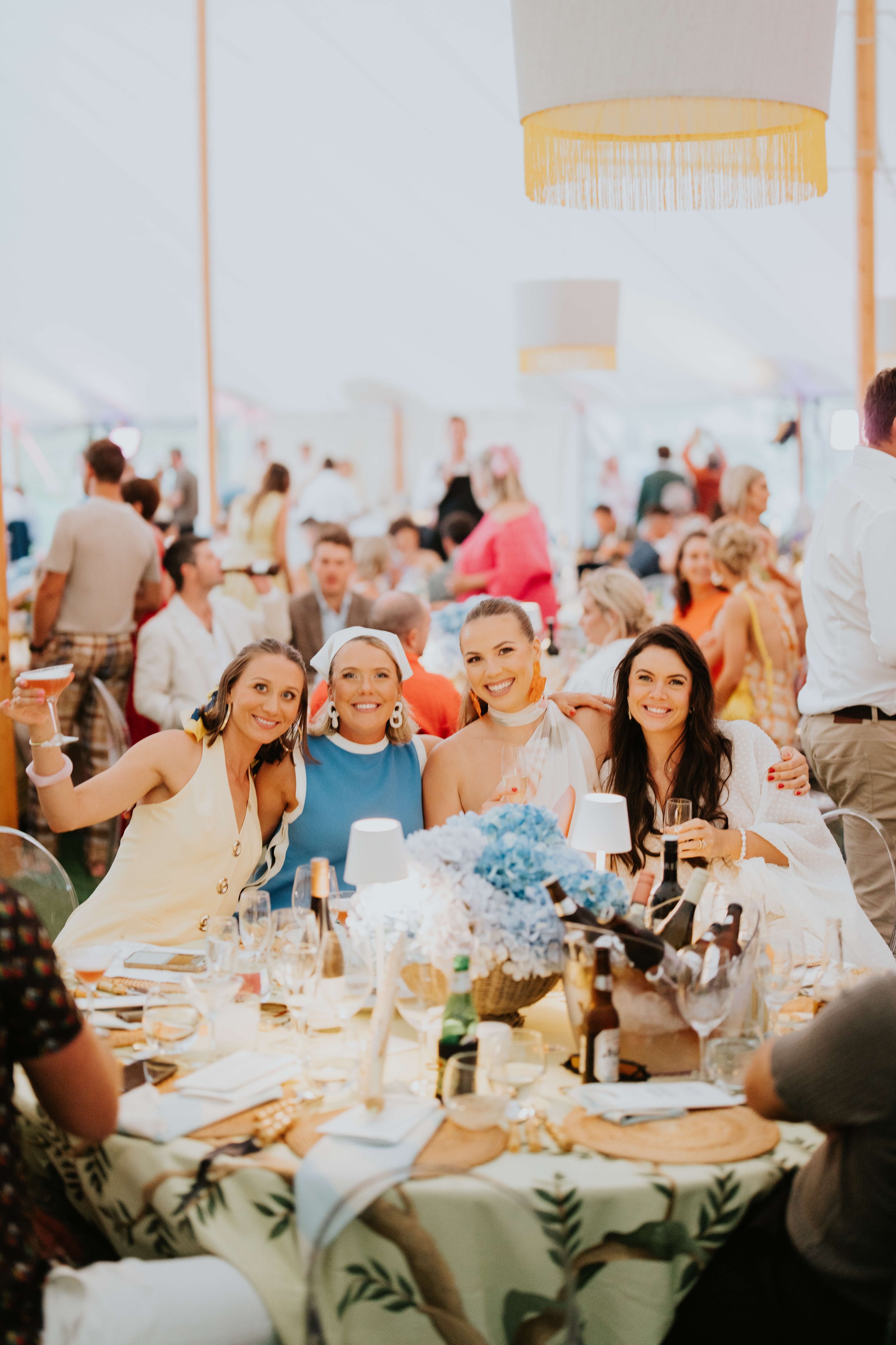A group of four women smiling and posing together at a table during an event. The table is decorated with blue hydrangeas, wine bottles, and glasses. They are in a well-lit, crowded indoor space with other attendees in the background.
