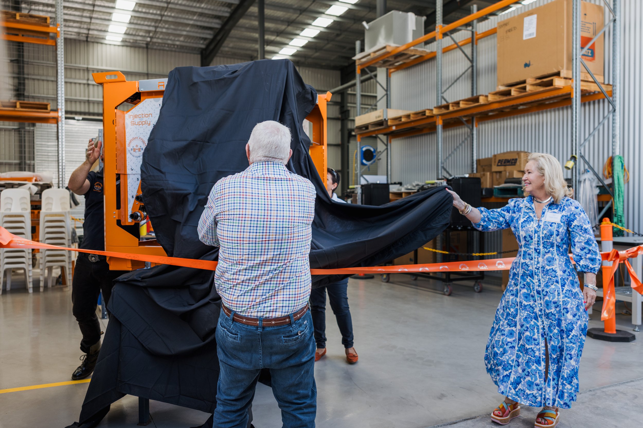 Group unveiling a machine in a warehouse setting, using a black sheet to cover it